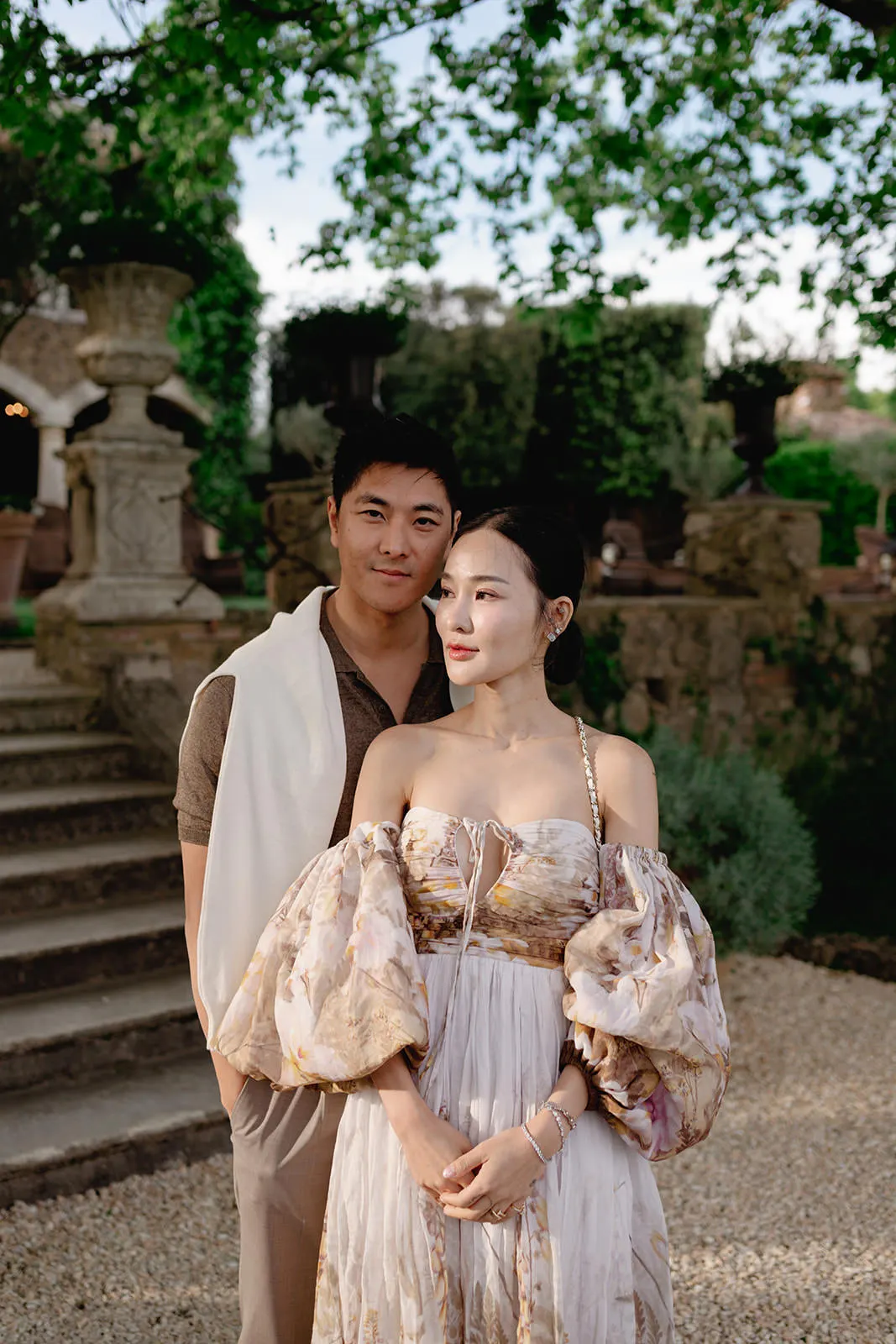 A man and woman stand outdoors in elegant clothing. The woman wears a light, off-the-shoulder dress with voluminous sleeves and floral patterns. The man wears a brown shirt with a white sweater draped over his shoulders. Green trees and stone steps are in the background. Tuscan Wedding Photographer