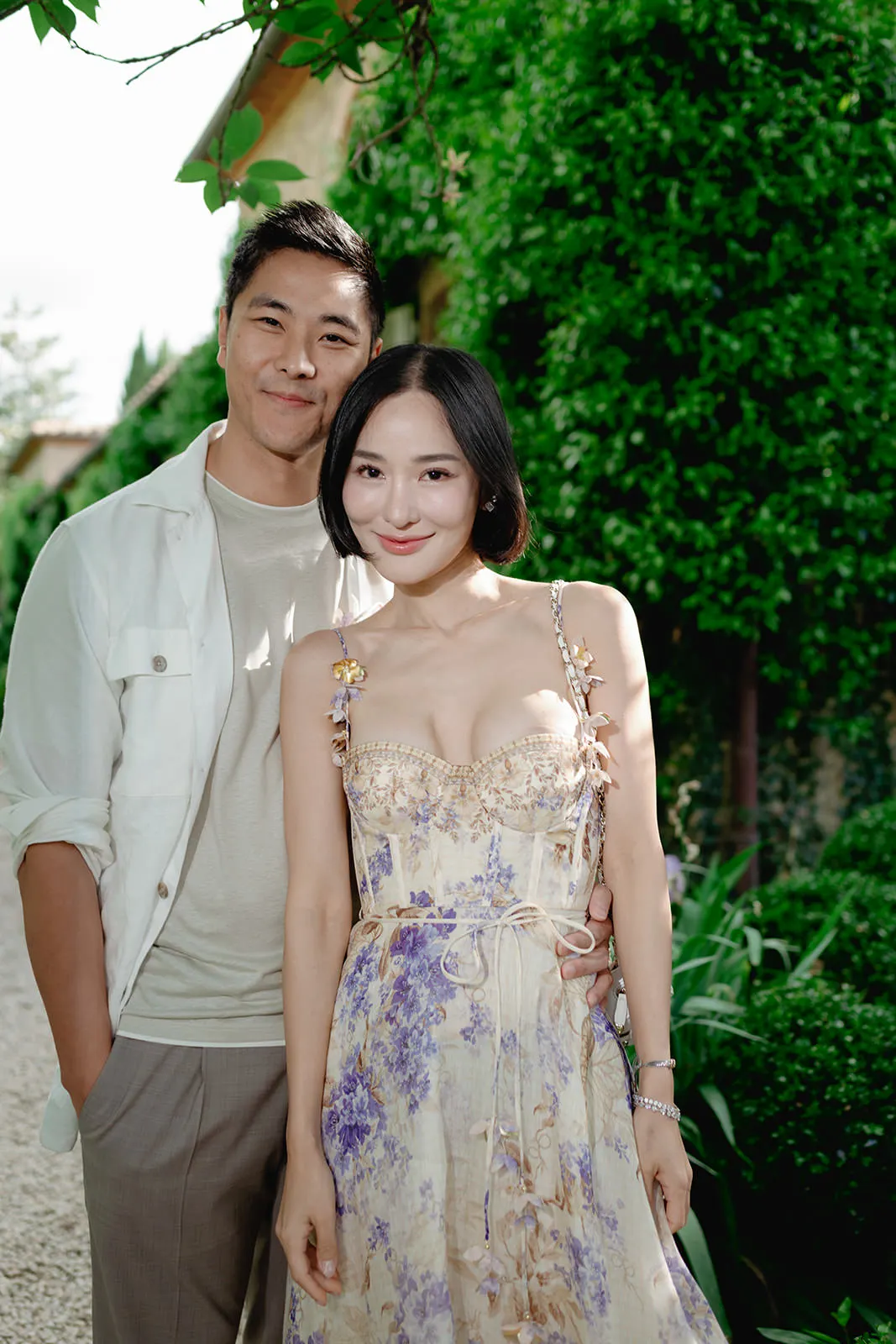 A smiling couple poses outdoors. The woman wears a floral, sleeveless dress with thin straps and a tied waist; the man stands slightly behind her, wearing a light shirt over a white tee and grey pants. Green foliage and sunlight fill the background. Tuscan Wedding Photographer