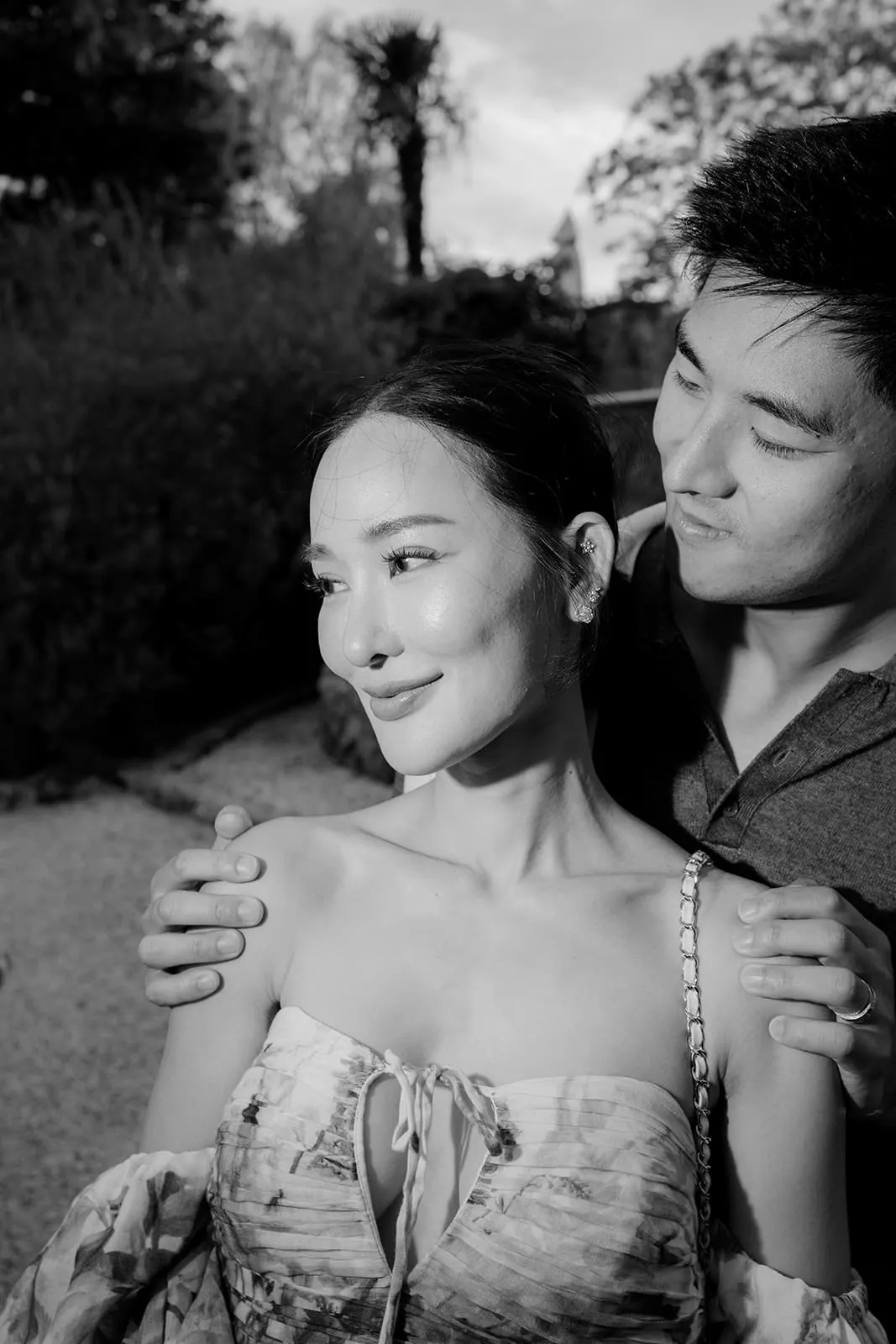 A black-and-white photo of a woman smiling slightly while a man stands behind her with his arms around her shoulders. They are outdoors, and both appear happy. The woman wears an off-shoulder dress; the man is in a collared shirt. Trees and bushes are in the background. Tuscan Wedding Photographer