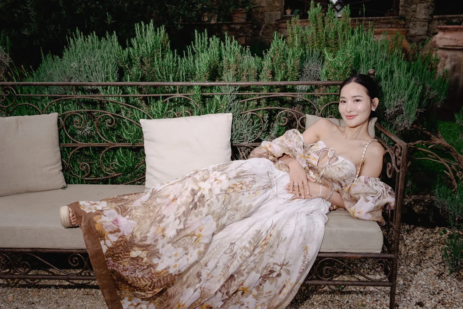 A woman in a long, flowing floral dress with puffed sleeves reclines on an ornate outdoor metal bench with beige cushions. She is smiling gently, surrounded by lush greenery and rustic stone walls in the background. Tuscan Wedding Photographer