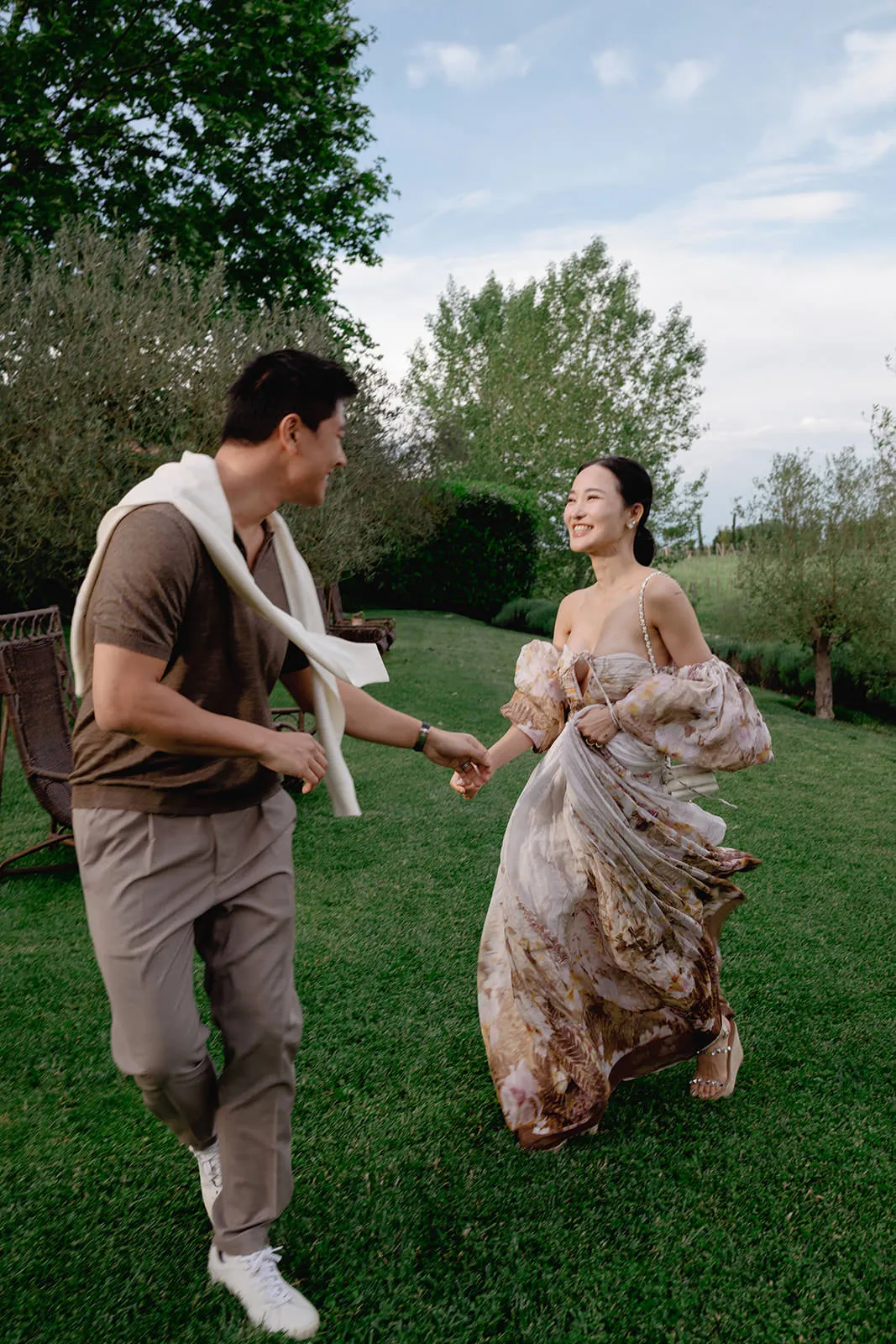 A smiling couple holding hands run on green grass in a garden. The man wears beige pants, a brown shirt, and a white sweater draped over his shoulders. The woman wears a flowing floral dress. Trees and shrubs line the background under a partly cloudy sky. Tuscan Wedding Photographer
