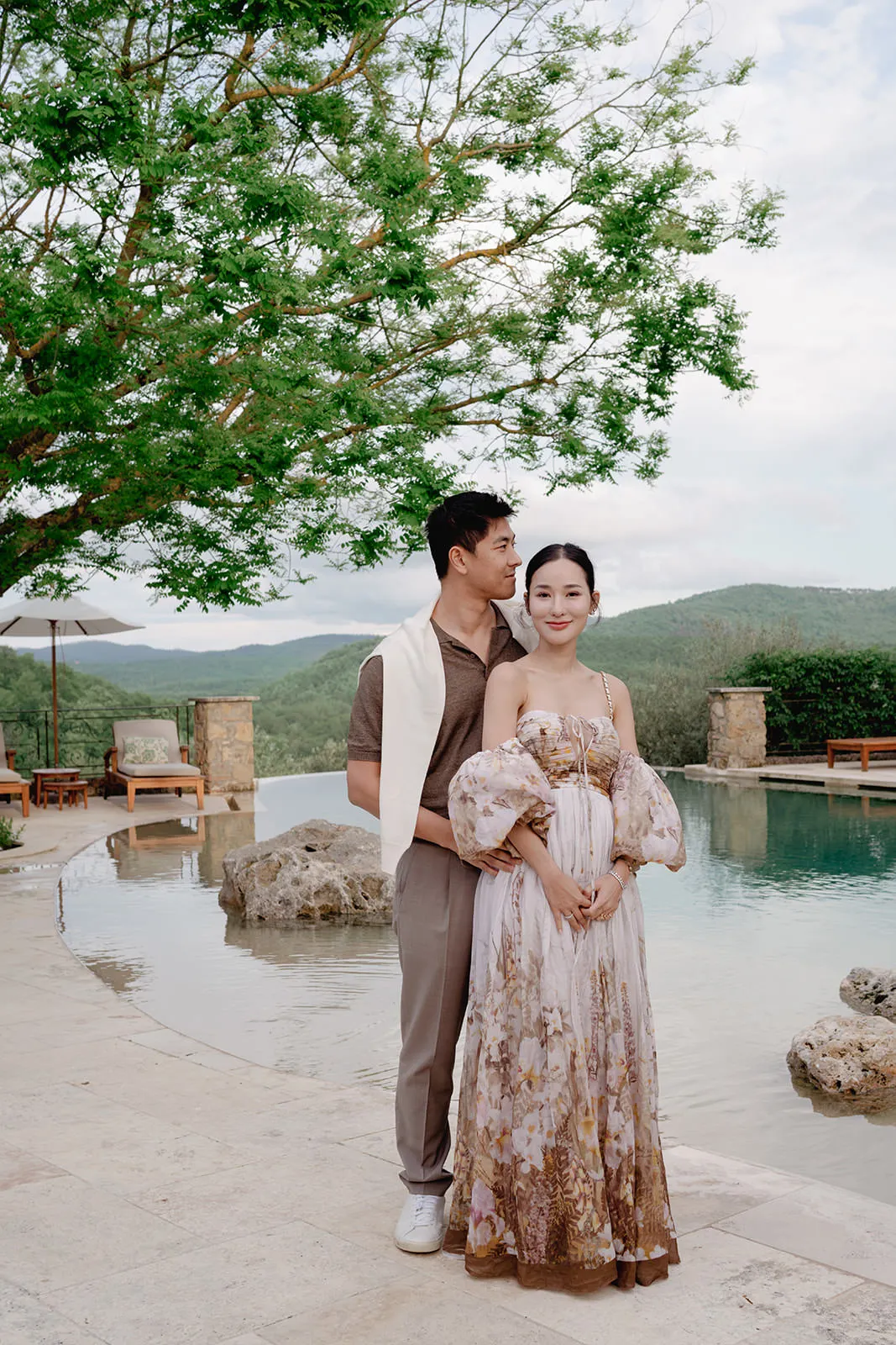 A couple stands by a serene, stone-edged pool. The man, in taupe pants, a brown shirt, and white sneakers, stands behind a woman in a flowing floral dress with puffy sleeves. Green trees and distant hills create a peaceful outdoor backdrop. Tuscan Wedding Photographer