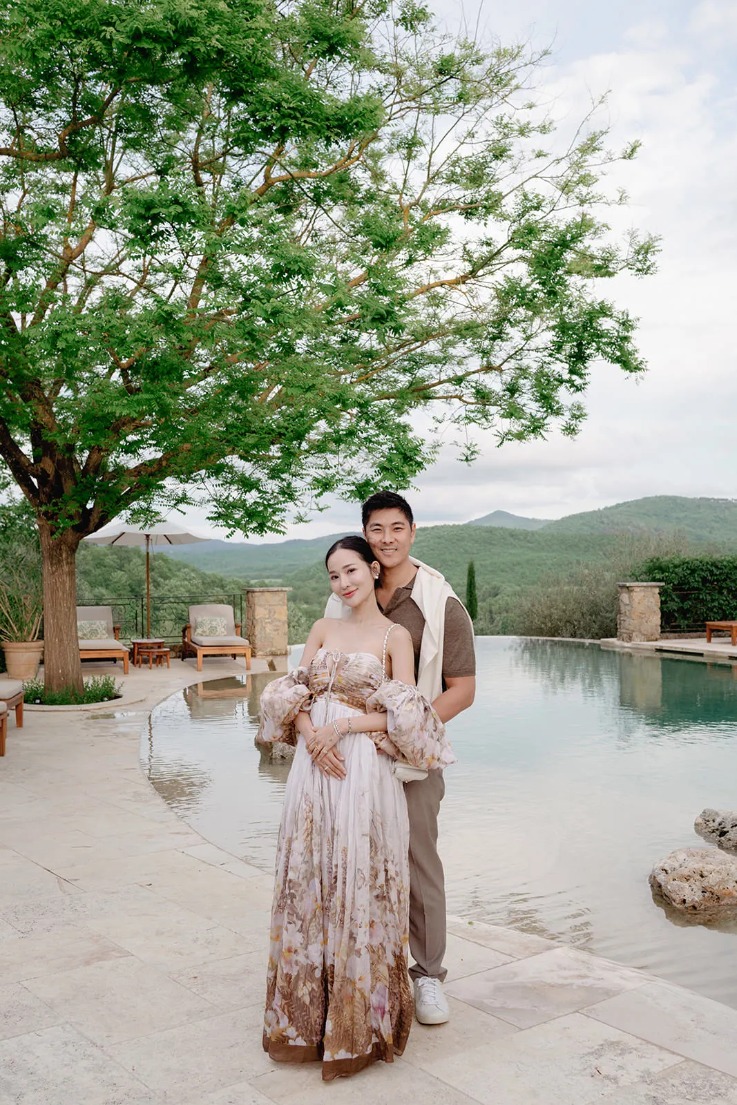 A couple poses by a serene infinity pool surrounded by stone tiles, plush chairs, and potted plants. The woman wears a flowing floral dress; the man stands behind her, hands on her waist, both smiling. A large green tree and rolling hills fill the background under a cloudy sky. Tuscan Wedding Photographer