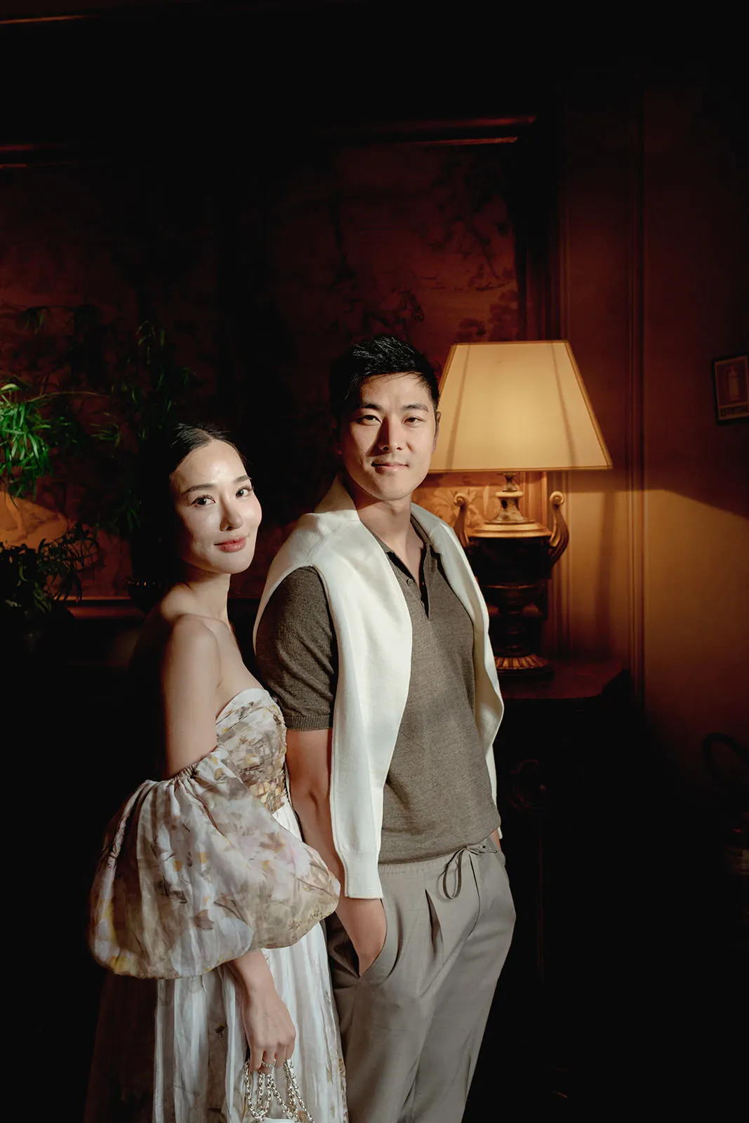 A woman in a floral, off-shoulder dress stands beside a man wearing a grey polo, beige pants, and a white sweater draped over his shoulders. They both face the camera and smile softly in a warmly lit, elegant room with a lamp and plants in the background. Tuscan Wedding Photographer