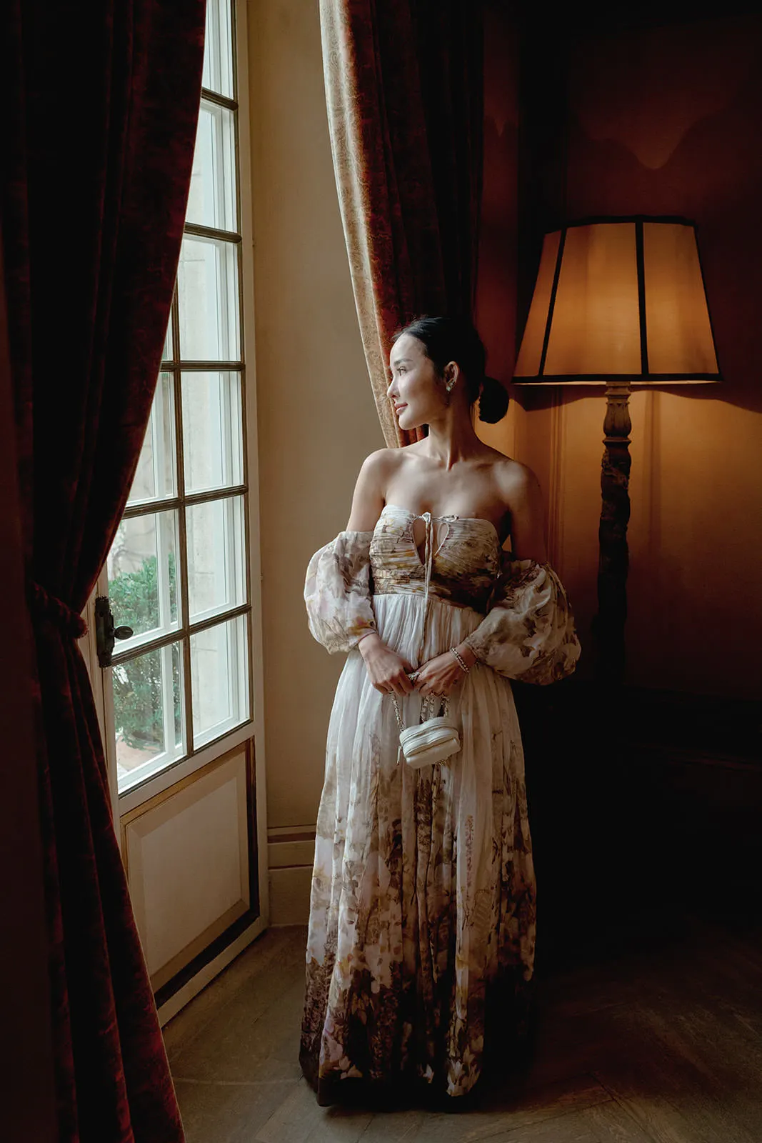 A woman in an off-the-shoulder, flowing floral gown stands by a large window with light streaming in. She holds a small white purse and looks outside, with her dark hair pulled back. A tall lamp with a beige shade glows in the softly lit room behind her. Tuscan Wedding Photographer