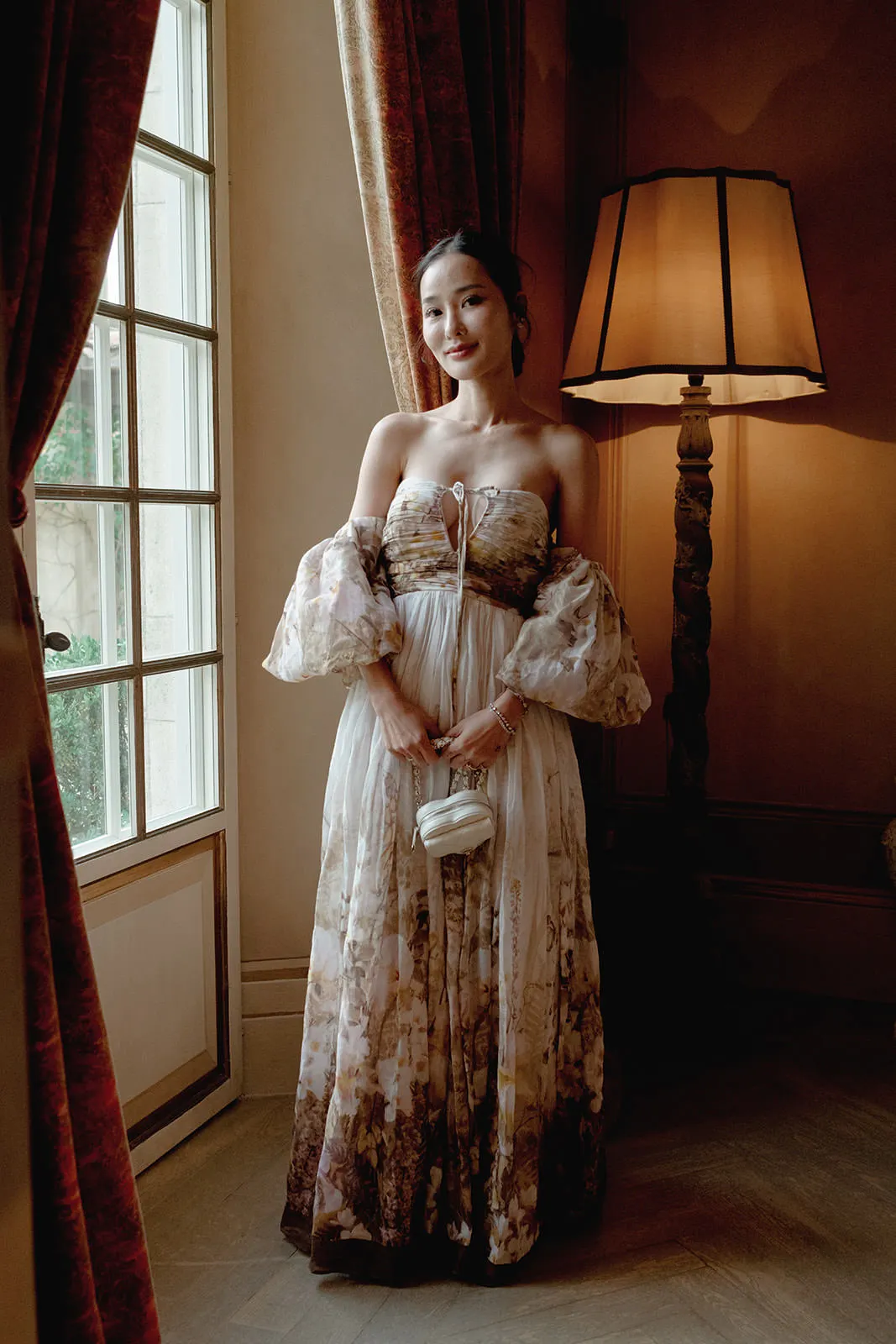 A woman stands indoors near a tall lamp and window, wearing an off-the-shoulder, flowing floral gown with sheer puff sleeves. She holds a small white purse with both hands and smiles softly. Warm light and classic decor create an elegant atmosphere. Tuscan Wedding Photographer
