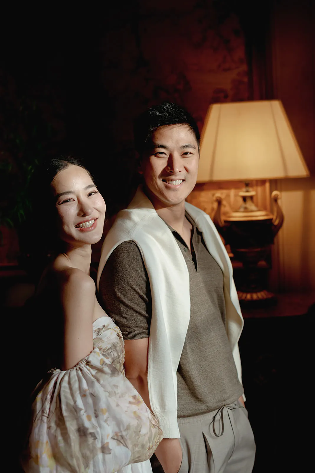 A smiling woman in a floral off-shoulder dress stands beside a smiling man in neutral-toned casual clothes with a white sweater draped over his shoulders. They pose together indoors next to a large ornate lamp emitting warm light. Tuscan Wedding Photographer
