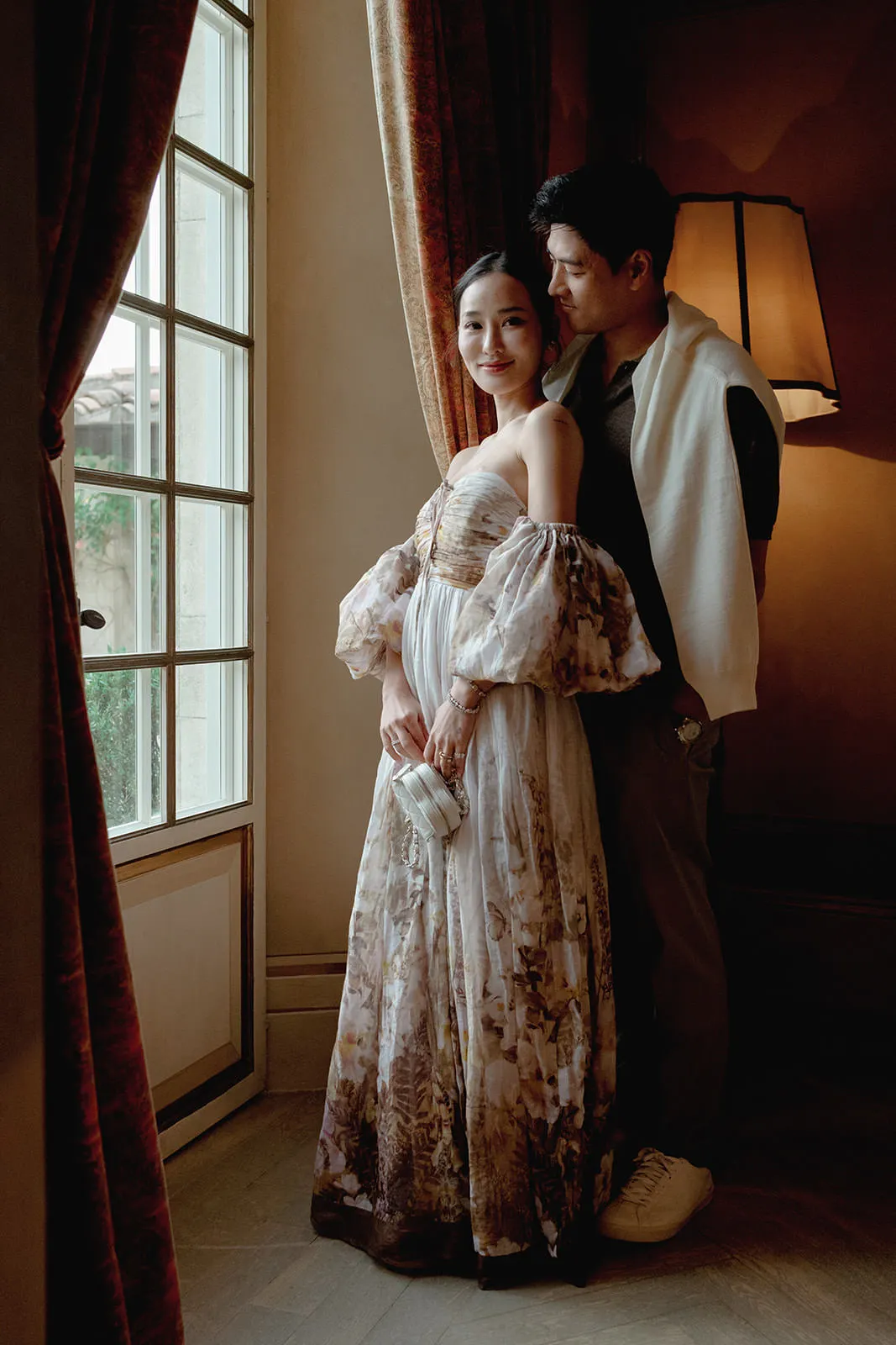 A woman in a flowing, off-shoulder floral dress stands by a window, holding a clutch and smiling softly. Behind her, a man in a dark outfit with a white sweater draped over his shoulders stands close, gazing at her. Warm indoor lighting creates a cozy atmosphere. Tuscan Wedding Photographer