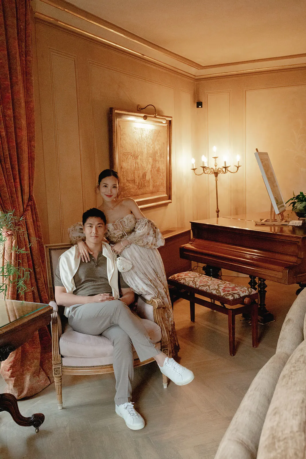 A man in casual clothes sits on a chair, while a woman in an elegant off-shoulder gown stands behind him with her hand on his shoulder. They are in a warmly lit, luxurious room with a grand piano, ornate furniture, gold curtains, and a framed painting. Tuscan Wedding Photographer