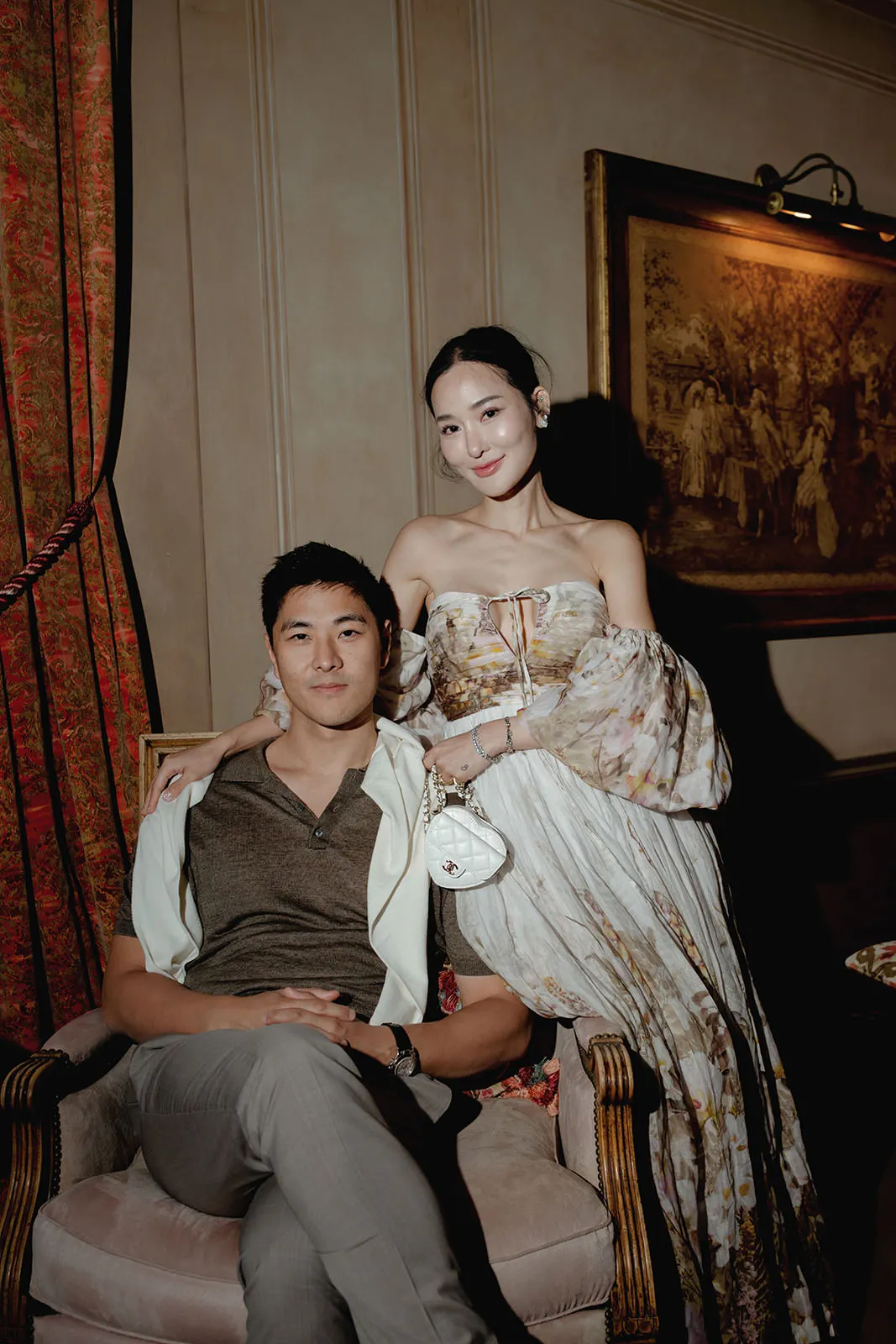 A man sits in a vintage armchair with his legs crossed, wearing a brown shirt and light pants. A woman in a flowing, floral off-the-shoulder dress stands beside him, hand on his shoulder. They pose in an elegant, dimly lit room with ornate curtains and framed art. Tuscan Wedding Photographer