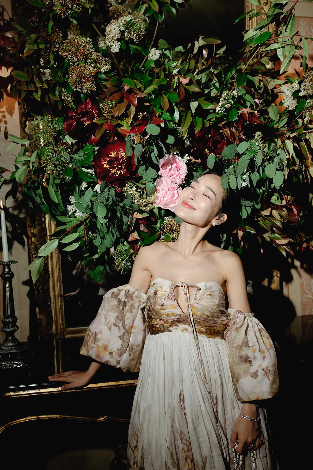 A woman in a floral, off-shoulder dress stands indoors, eyes closed and smiling slightly, her face near a large, lush bouquet of dark red, white, and pink flowers with abundant green foliage. Soft, warm lighting enhances the romantic, elegant atmosphere. Tuscan Wedding Photographer