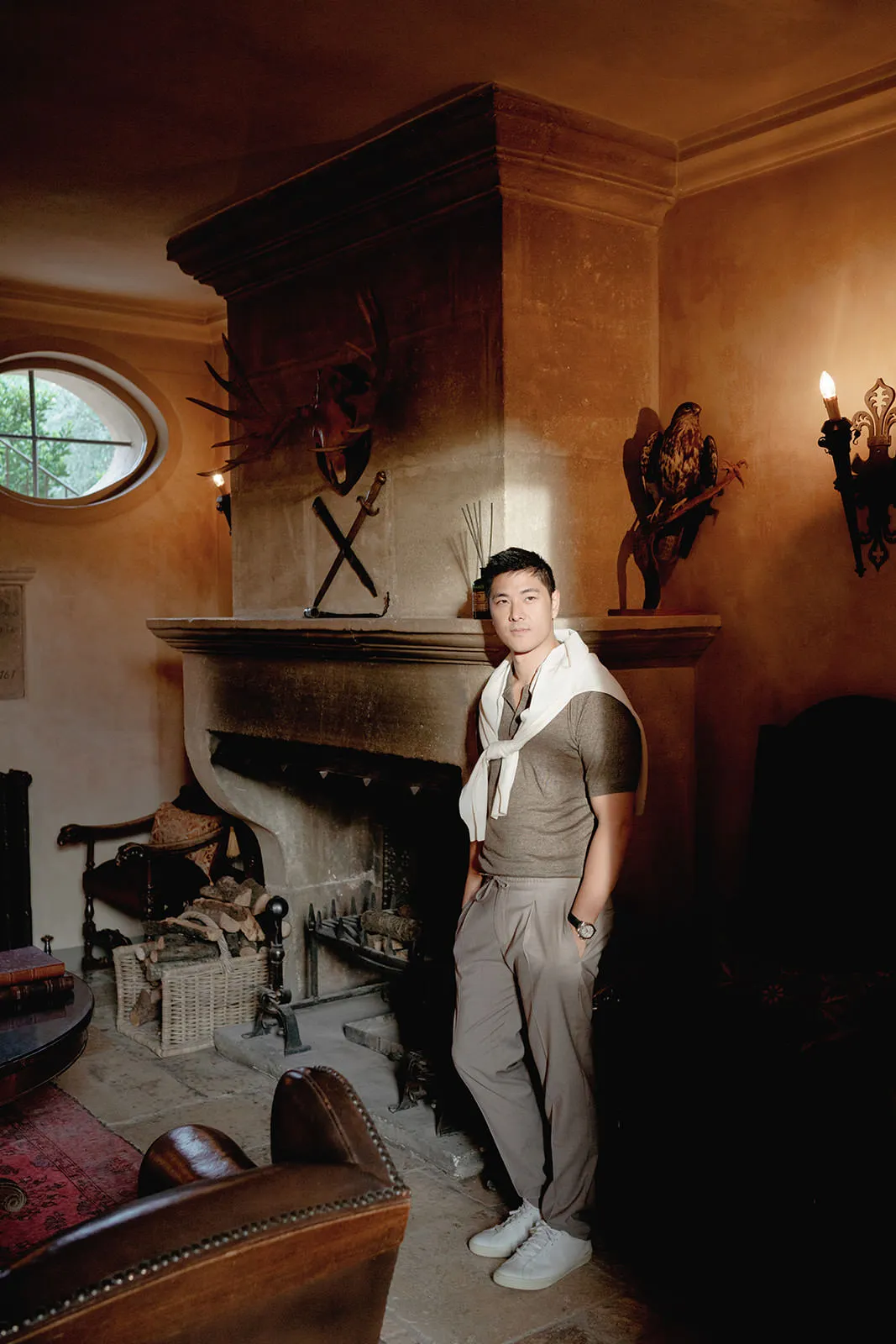 A man stands casually by a large stone fireplace in a warmly lit, rustic room. He wears light trousers, a brown shirt, white sneakers, and has a white sweater draped over his shoulders. Antique decor, armchairs, and animal wall mounts enhance the cozy atmosphere. Tuscan Wedding Photographer