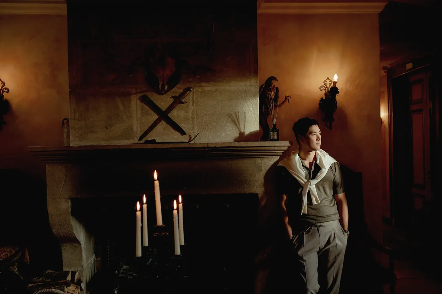 A man stands beside a large stone fireplace with five lit candles. He wears a sweater draped over his shoulders and gazes to the side. The room is dimly lit, with warm wall lights and antique decor including a statue, axes, and animal head above the mantle. Tuscan Wedding Photographer