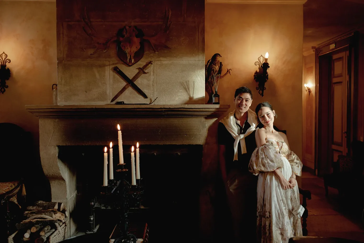 A couple in vintage attire stands by a stone fireplace adorned with candles, a skull, crossed tools, and a bird statue. The warmly lit room features ornate wall sconces, wood paneling, and a cozy, elegant atmosphere. The couple smiles gently. Tuscan Wedding Photographer