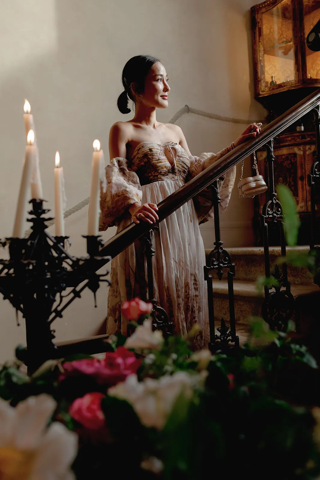 A woman in an ornate, off-shoulder gown stands on a staircase, holding the railing. She is smiling softly and looking to her right. In the foreground, lit candles and pink and white flowers are visible, with antique decor in the background. Tuscan Wedding Photographer