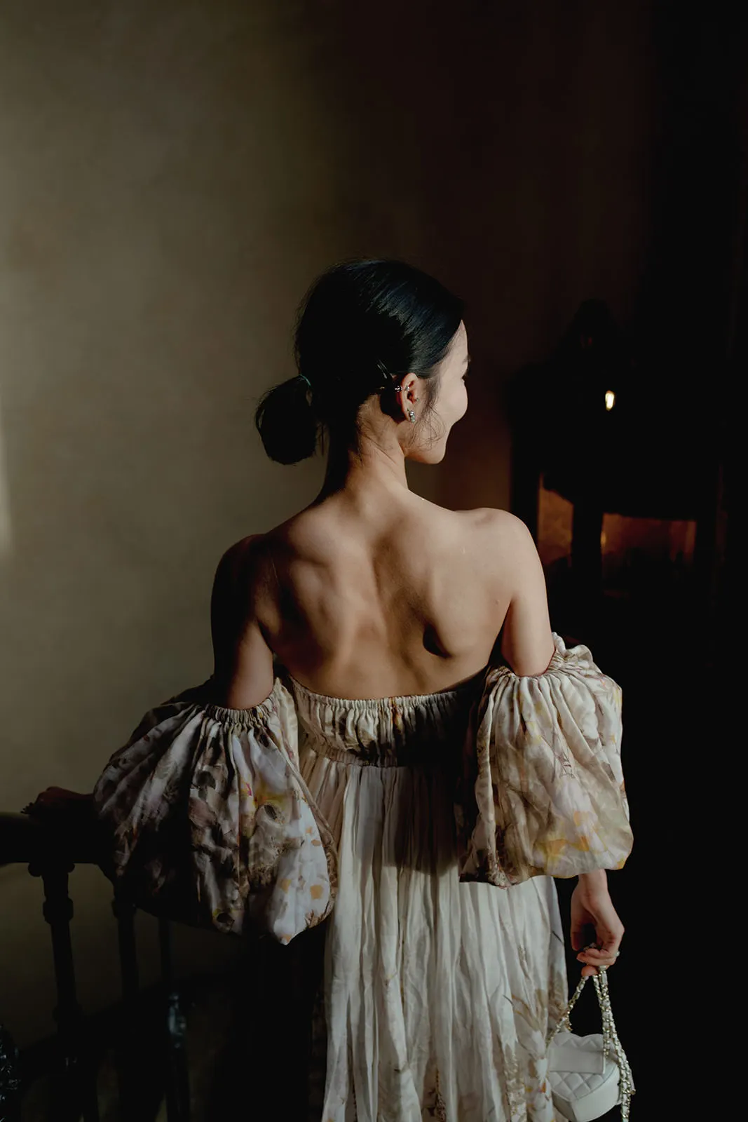 A woman with dark hair in a low bun stands in a dimly lit room, seen from behind. She wears a flowing off-the-shoulder dress with voluminous, patterned sleeves, holding a small white purse in her right hand. Light softly highlights her bare back. Tuscan Wedding Photographer