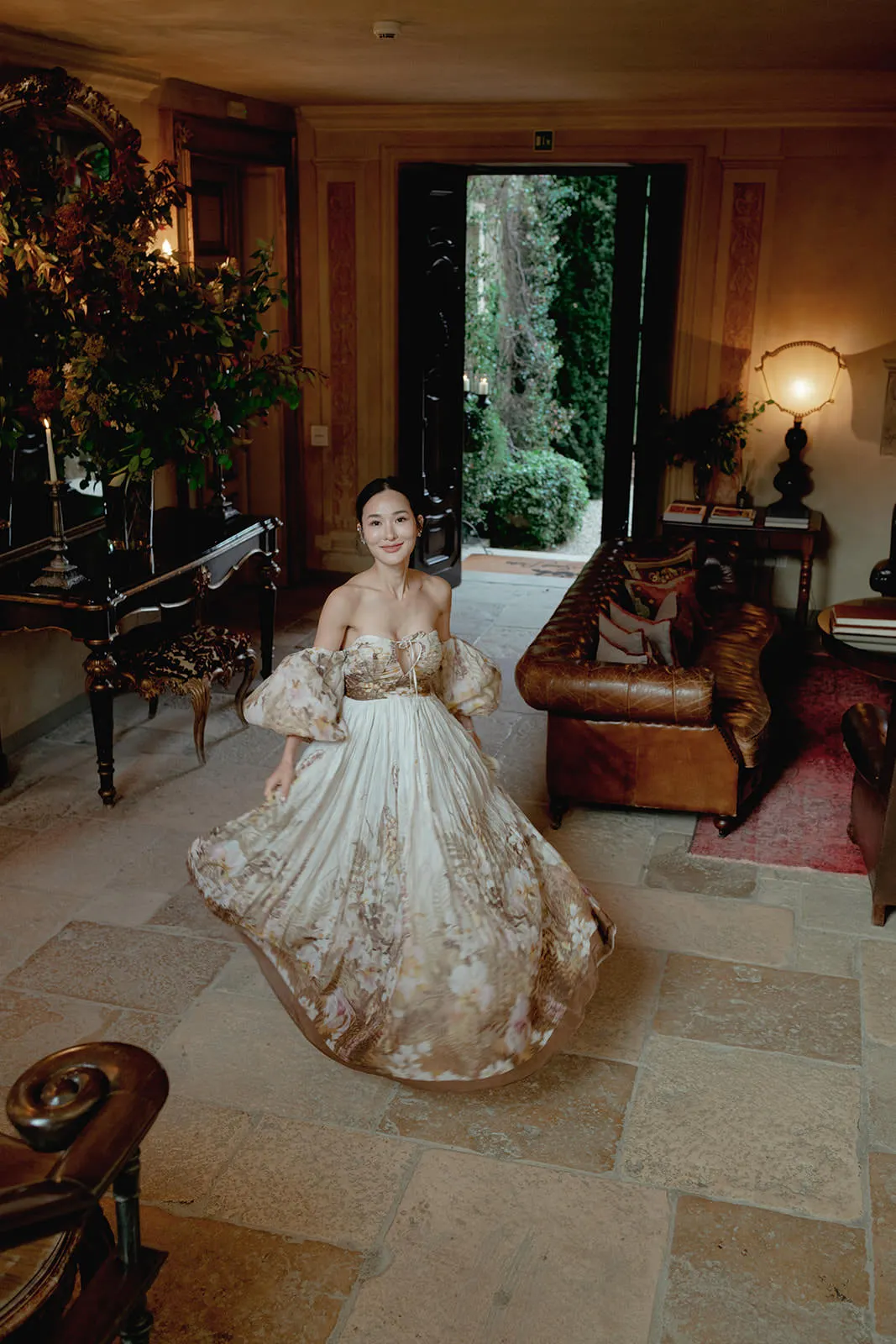 A woman in a flowing, off-shoulder floral gown twirls in an elegant, warmly lit room with stone floors, a grand mirror, leather sofas, a piano, and an open door revealing a green garden outside. Tuscan Wedding Photographer
