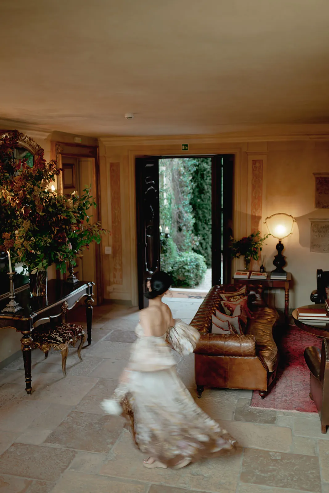 A blurred woman in a flowing dress walks through an elegant, vintage living room with a leather sofa, ornate mirror, floral arrangement, and open door revealing a lush garden outside. Warm lighting and classic decor create a cozy, sophisticated atmosphere. Tuscan Wedding Photographer