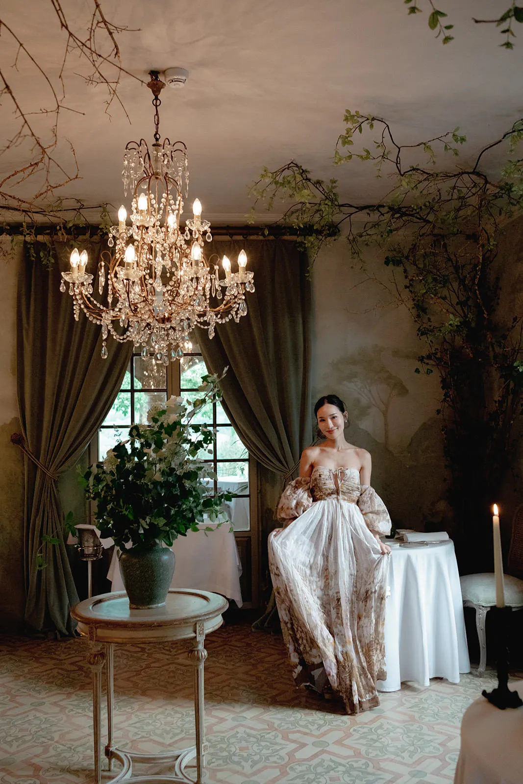 A woman in an elegant, flowing floral gown stands by a table in a vintage-style room with leafy branches, draped curtains, and a large window. A crystal chandelier hangs overhead, and a potted plant sits on a round table in the foreground. Tuscan Wedding Photographer