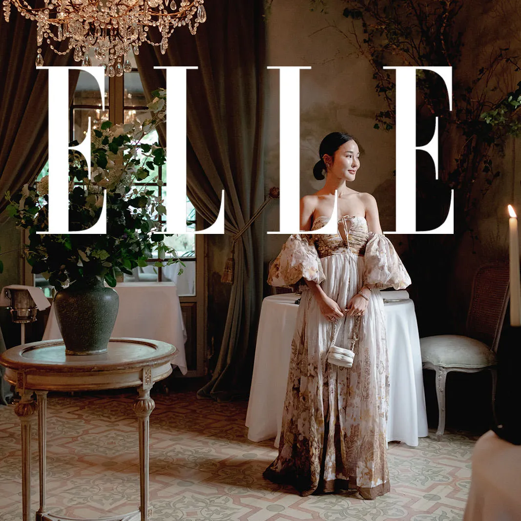 A woman in a flowing, off-shoulder floral gown stands in a stylish, elegant room with a chandelier and candlelight—reminiscent of engagement photography in Tuscany. Large white ELLE letters are superimposed, evoking a magazine cover or editorial. Tuscan Wedding Photographer