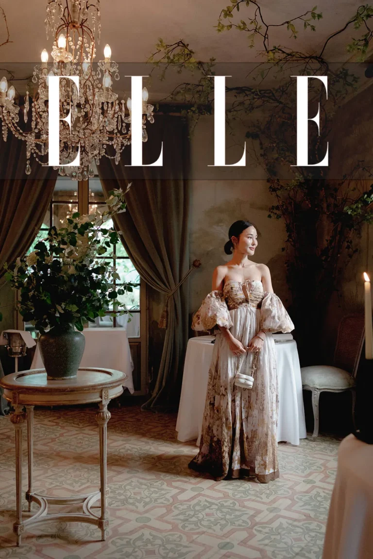 A woman in an elegant, floral off-shoulder gown stands in a vintage, ornate room with lush greenery, chandeliers, and candlelight. Sunlight filters through draped curtains. The word ELLE is boldly overlaid at the top of the image. Tuscan Wedding Photographer
