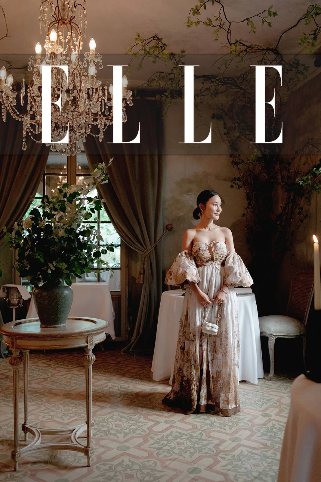 A woman in an elegant, floral off-shoulder gown stands in a vintage, ornate room with lush greenery, chandeliers, and candlelight. Sunlight filters through draped curtains. The word ELLE is boldly overlaid at the top of the image. Tuscan Wedding Photographer
