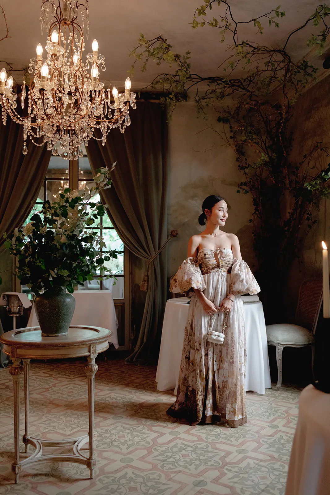 A woman in an elegant, flowing gown stands in a dimly lit, ornate room with a chandelier and classical decor—an enchanting scene perfect for a wedding photographer Tuscany dreams of capturing. Tuscan Wedding Photographer