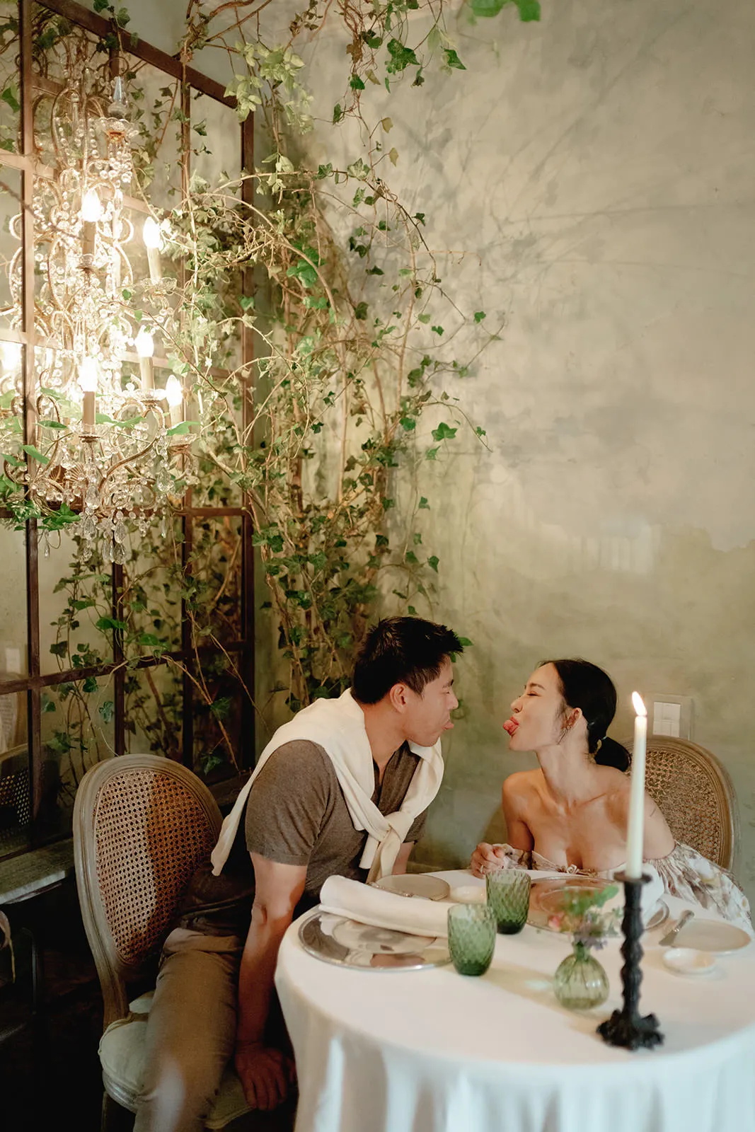 A couple sits at an elegant table set for two, playfully leaning toward each other as the woman sticks out her tongue. They are surrounded by lush greenery, ornate lighting, and a softly lit romantic atmosphere with a tall candle on the table. Tuscan Wedding Photographer