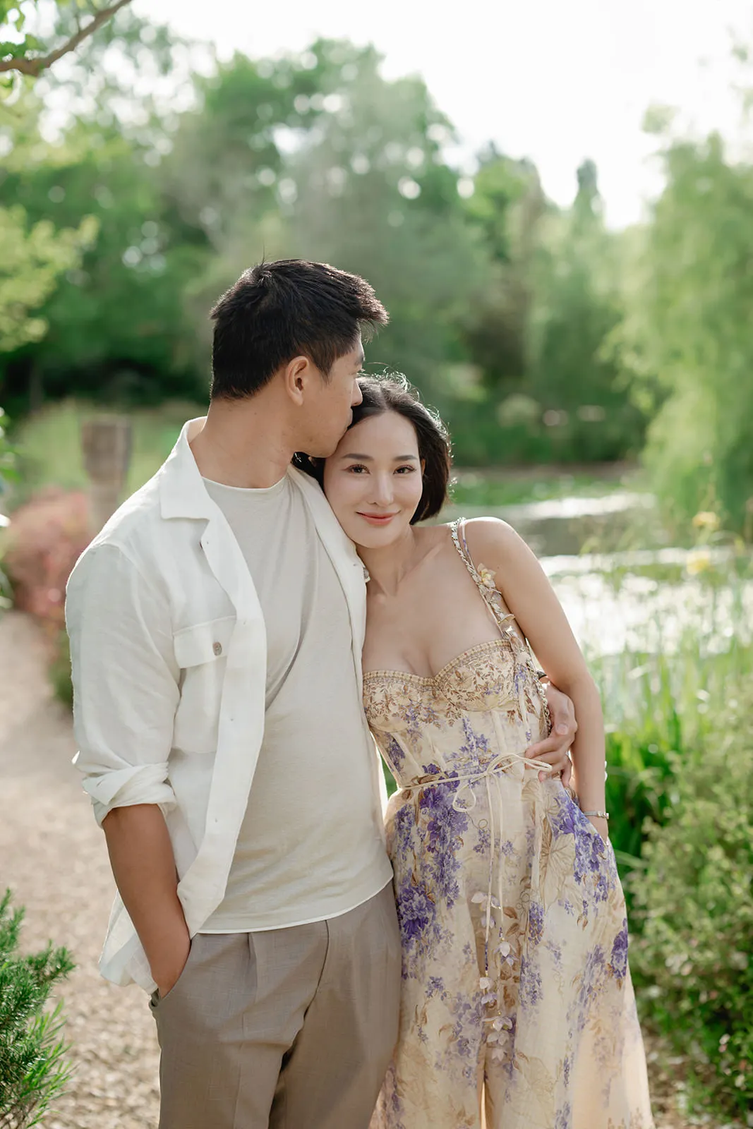 A couple poses outdoors by a pond and greenery. The man wears a light shirt and pants, embracing the woman, who wears a light floral dress. She smiles at the camera while he kisses her head. The scene is bright and serene, with lush green foliage in the background. Tuscan Wedding Photographer