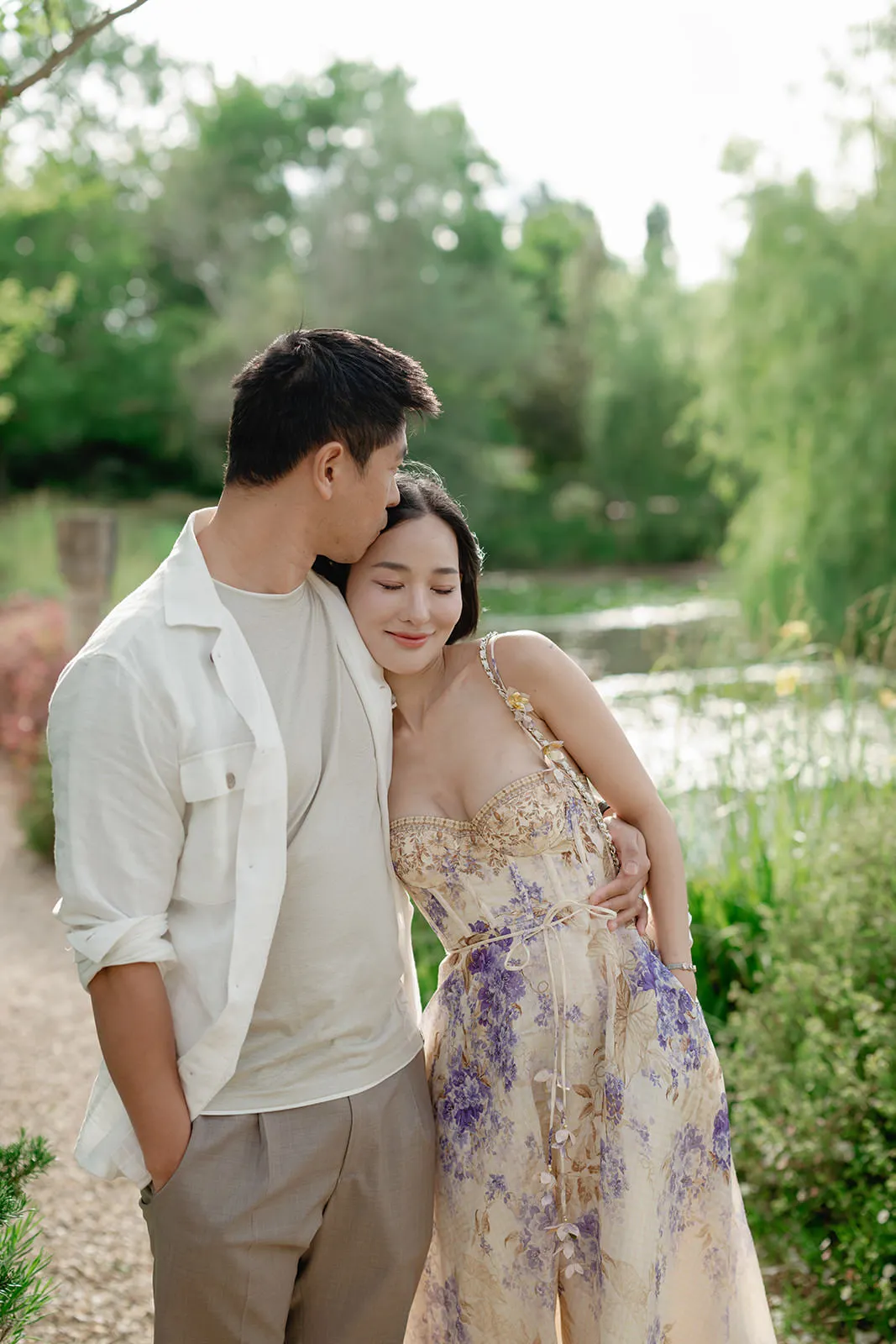 A couple stands outdoors by a pond and lush greenery. The man, in a light shirt and trousers, gently kisses the forehead of the smiling woman who wears a light, floral dress with purple accents. She closes her eyes and leans affectionately into him. Tuscan Wedding Photographer