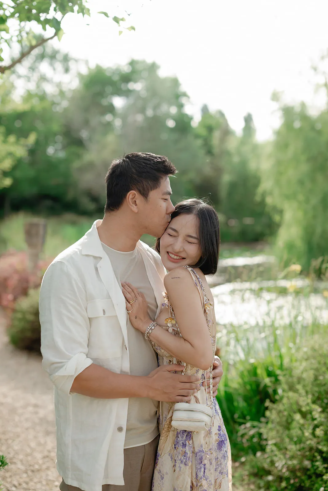 A man kisses a smiling woman on the forehead as they embrace outdoors. Both appear happy; the woman holds his chest. They stand on a path beside lush green plants and a pond, with sunlight filtering through trees in the background. Tuscan Wedding Photographer