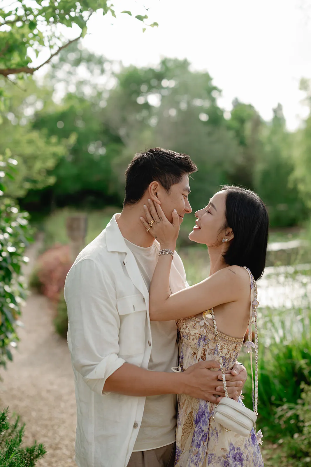 A couple stands outdoors on a garden path, embracing and smiling at each other. The woman holds the man’s face lovingly with one hand, while he holds her waist. Lush greenery and soft sunlight fill the peaceful background. Tuscan Wedding Photographer