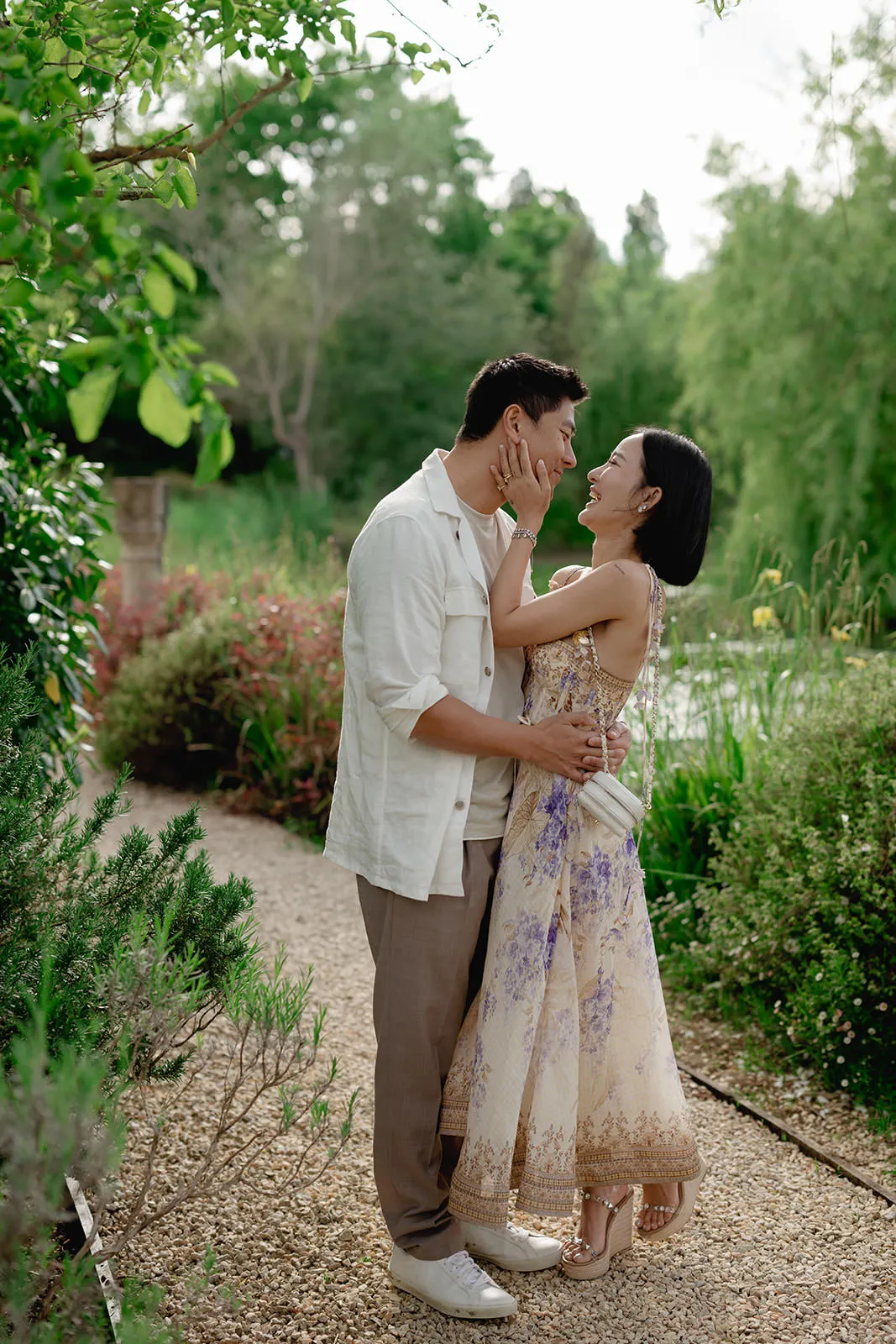A couple stands on a garden path, embracing affectionately. The woman, in a floral dress and sandals, gently holds the mans face. He wears a white shirt, tan pants, and white shoes. Lush greenery and colorful plants surround them, creating a romantic outdoor setting. Tuscan Wedding Photographer
