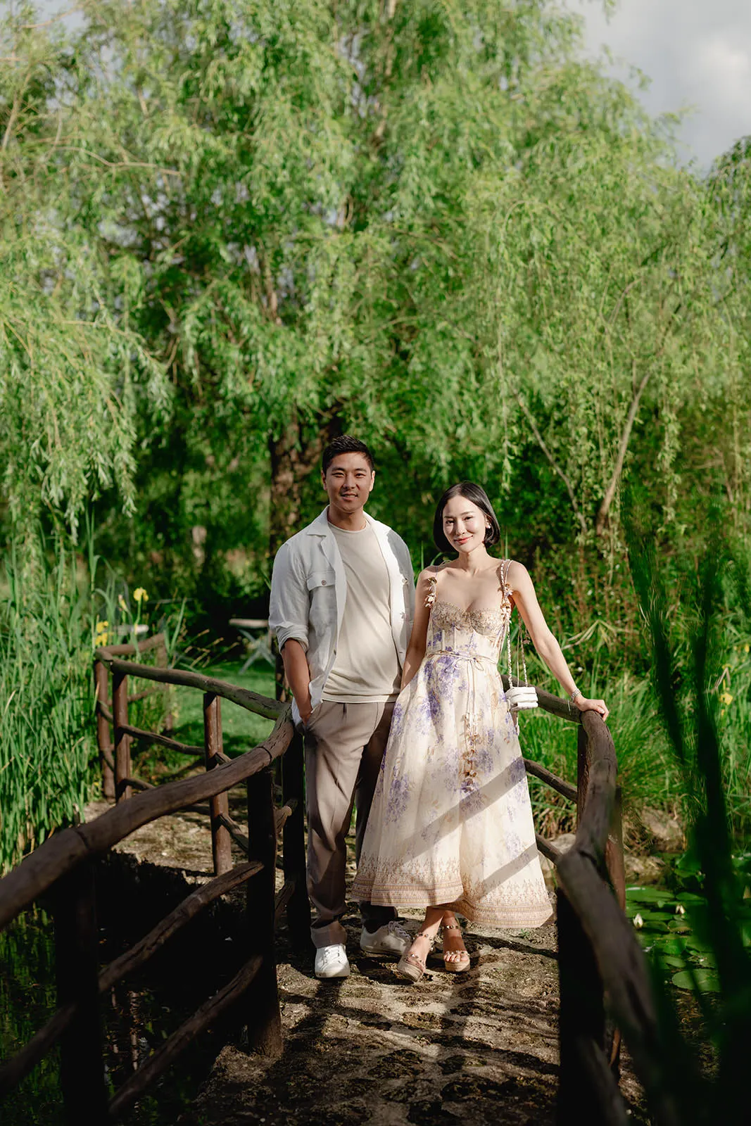 A smiling man and woman stand side by side on a rustic wooden bridge in a lush, green park. The woman wears a long, floral summer dress; the man wears light pants and a casual shirt. Tall trees and greenery surround them in the bright daylight. Tuscan Wedding Photographer