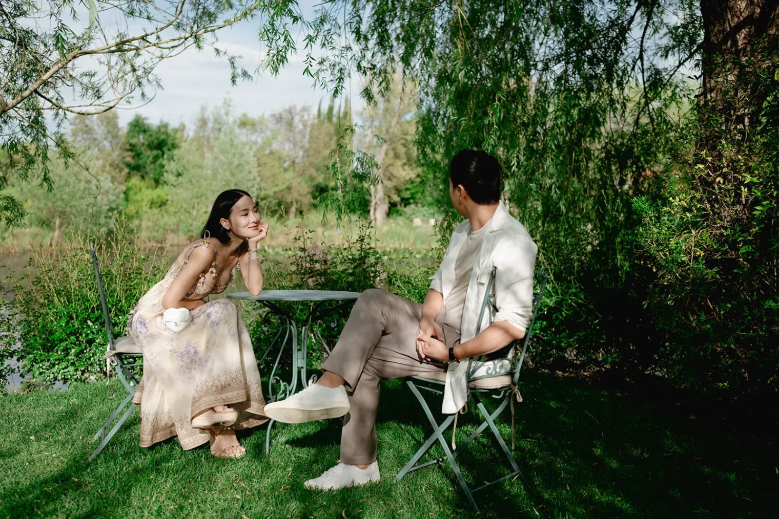 A woman in a light, floral dress and a man in a white shirt and khakis sit on metal chairs at a round table outdoors, surrounded by greenery and trees, with sunlight filtering through the leaves. The woman smiles, resting her chin on her hand. Tuscan Wedding Photographer