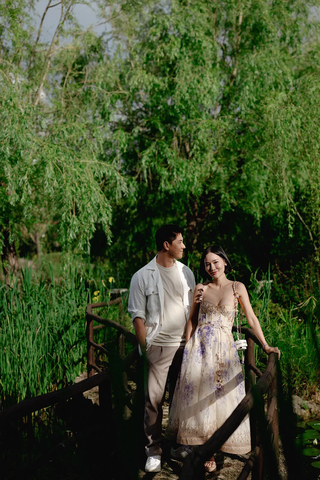 A couple stands on a wooden bridge surrounded by lush green foliage. The man wears a light jacket and trousers, while the woman, smiling and holding a phone, wears a light floral dress. Sunlight filters through the trees, creating a serene, romantic scene. Tuscan Wedding Photographer