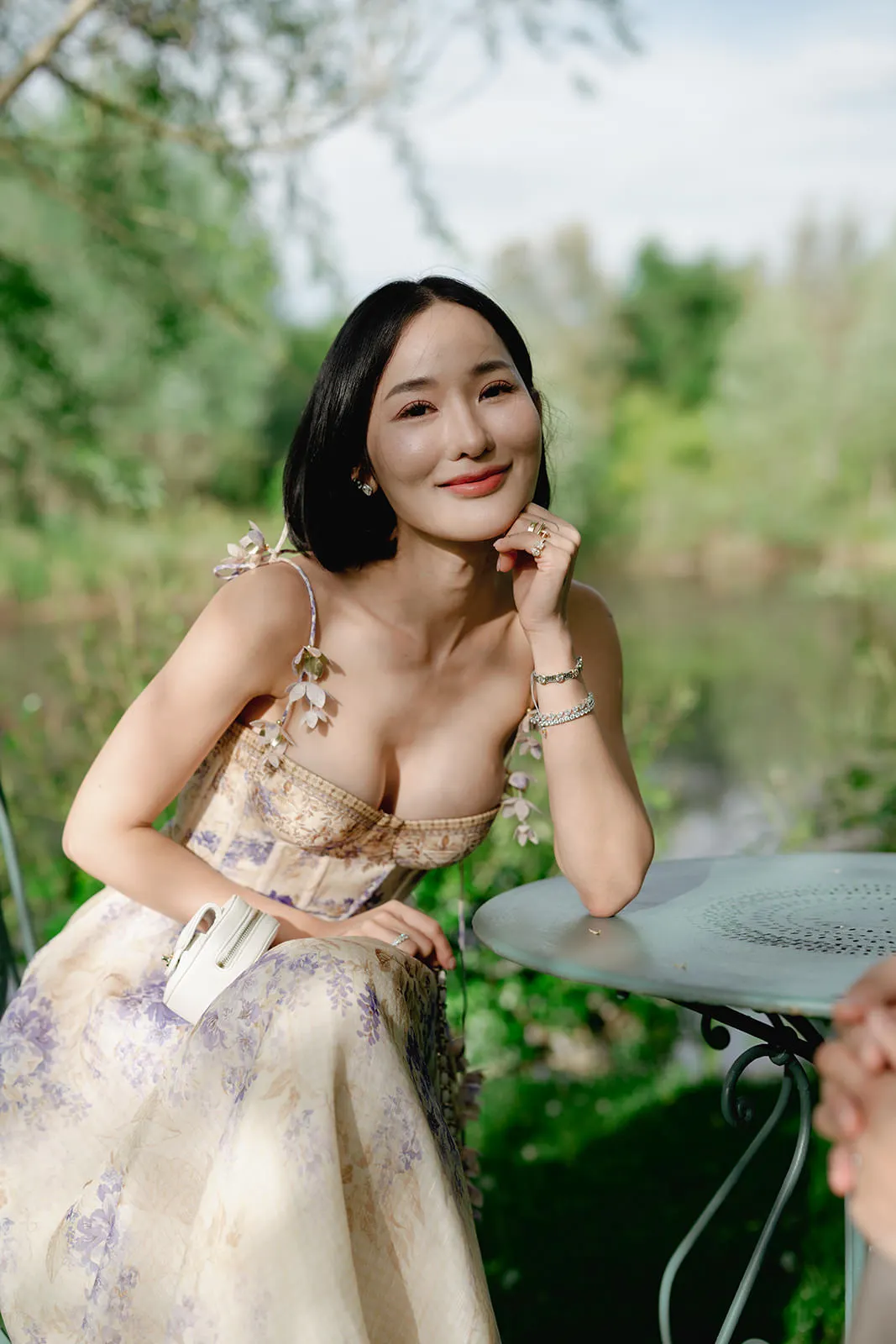 A woman with long black hair, wearing a floral, light-colored dress with spaghetti straps, sits outdoors at a round metal table. She smiles gently, resting her chin on her hand, with a blurred natural, green background behind her. Tuscan Wedding Photographer