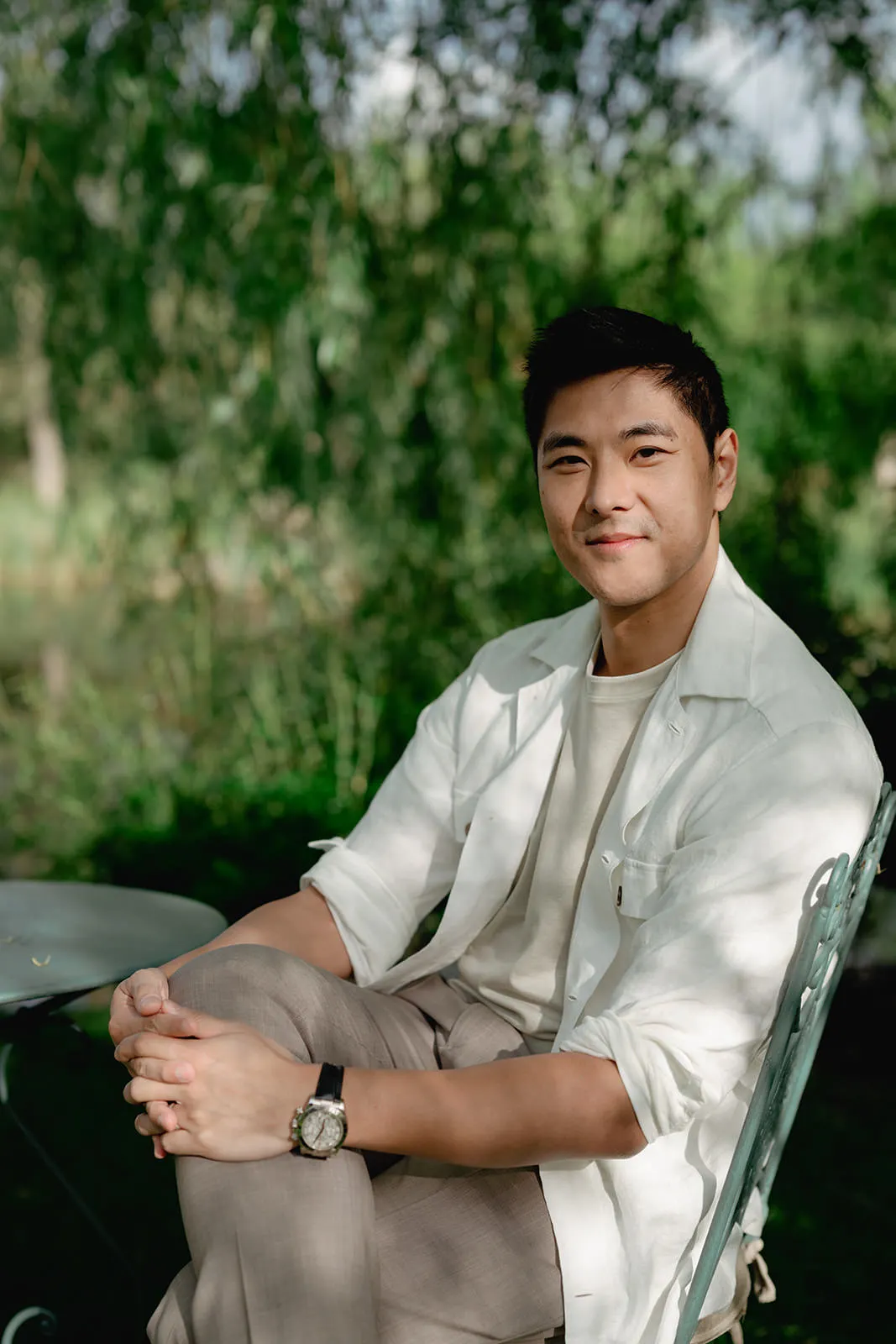 A man with short black hair, wearing a white shirt over a light t-shirt and beige pants, sits on an outdoor metal chair with his hands folded and legs crossed. He smiles softly, surrounded by blurred green trees and sunlight in the background. Tuscan Wedding Photographer