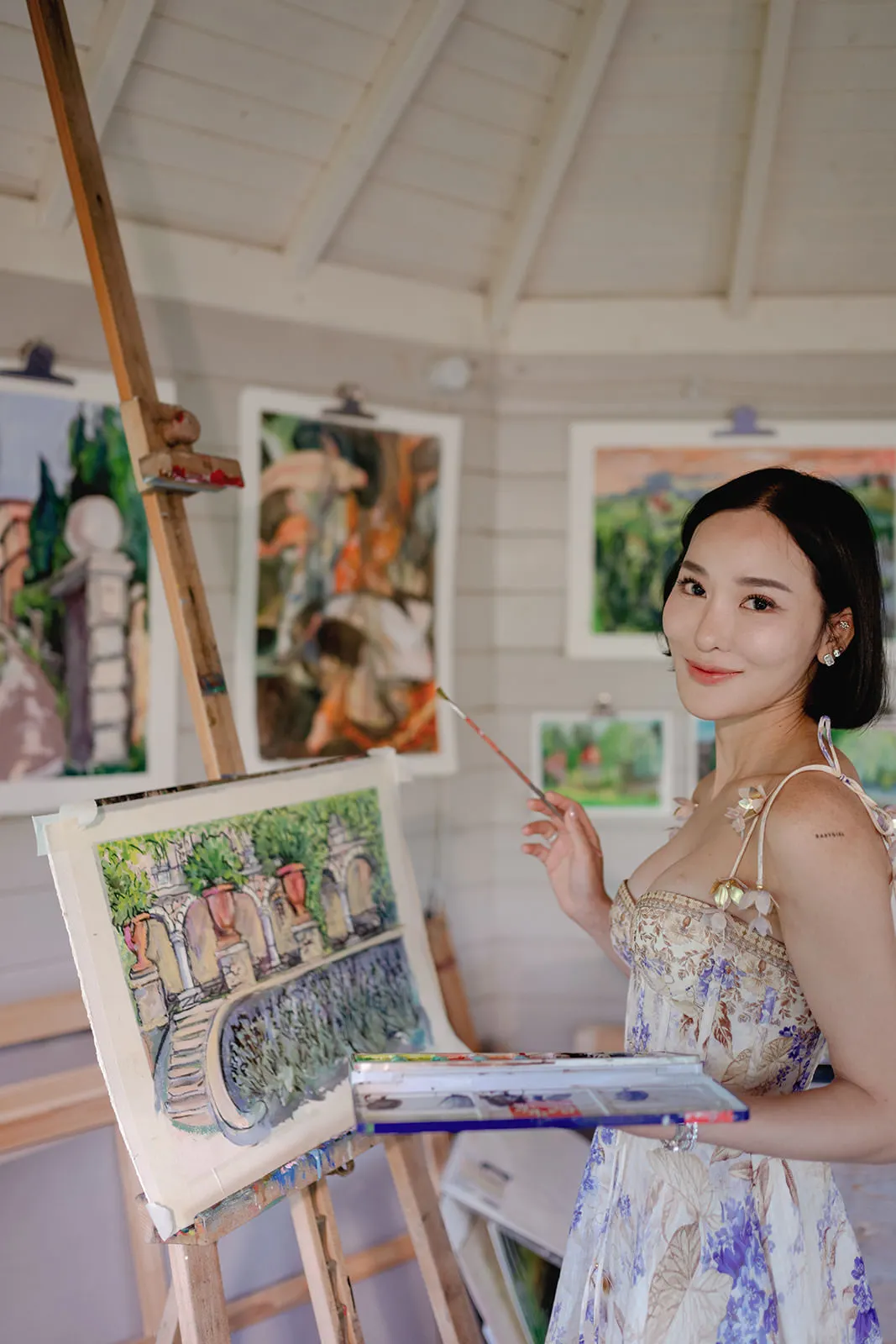 A woman in a floral dress stands in an art studio, smiling at the camera while painting a colorful landscape on a canvas. She holds a palette and brush, surrounded by finished paintings displayed on the walls of the bright, wooden room. Tuscan Wedding Photographer