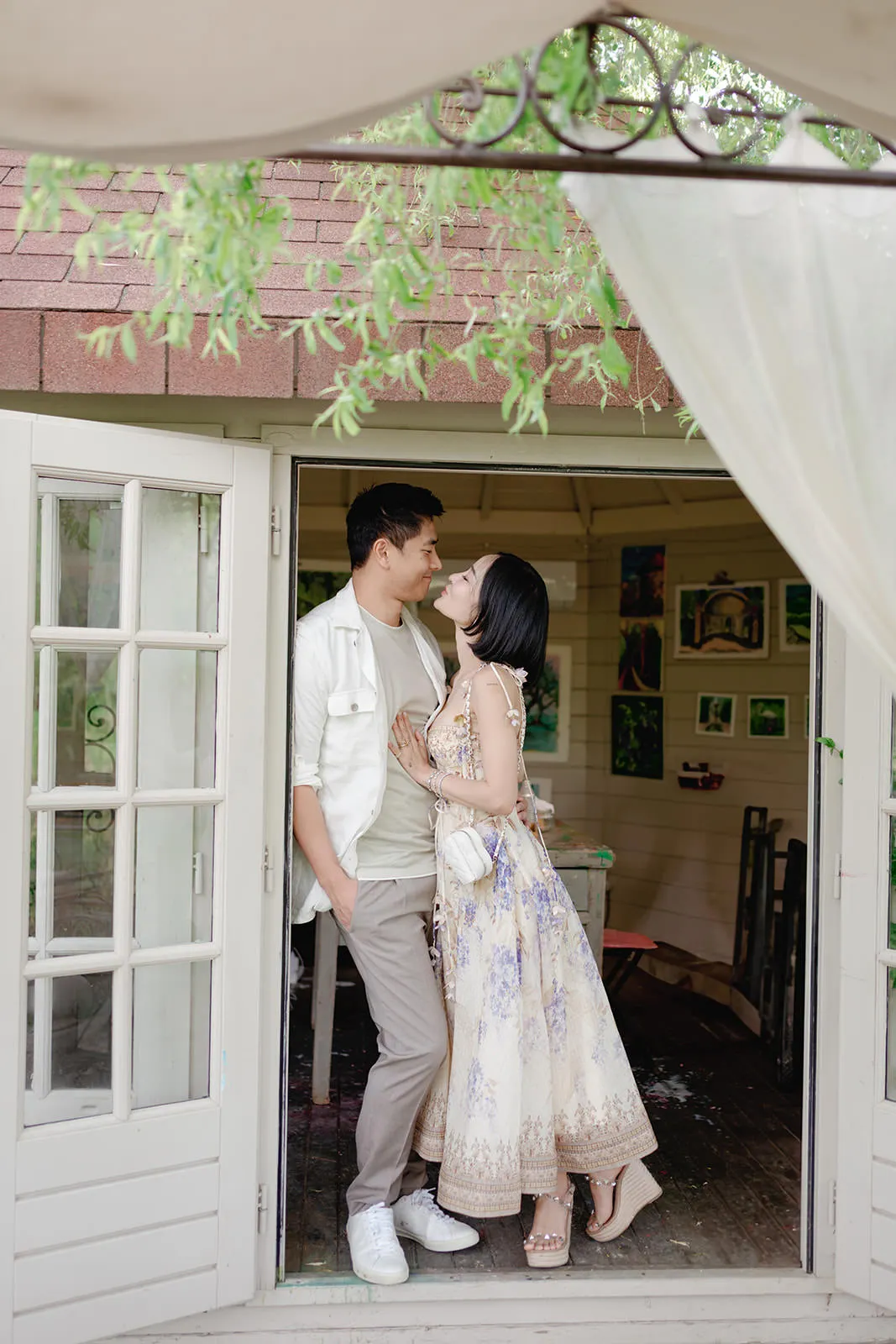 A couple stands in the doorway of a cozy, art-filled studio. The man in light casual clothes gazes at the woman, who wears a flowing floral dress and sandals. They smile warmly at each other under leafy branches and a sheer curtain, creating a romantic scene. Tuscan Wedding Photographer