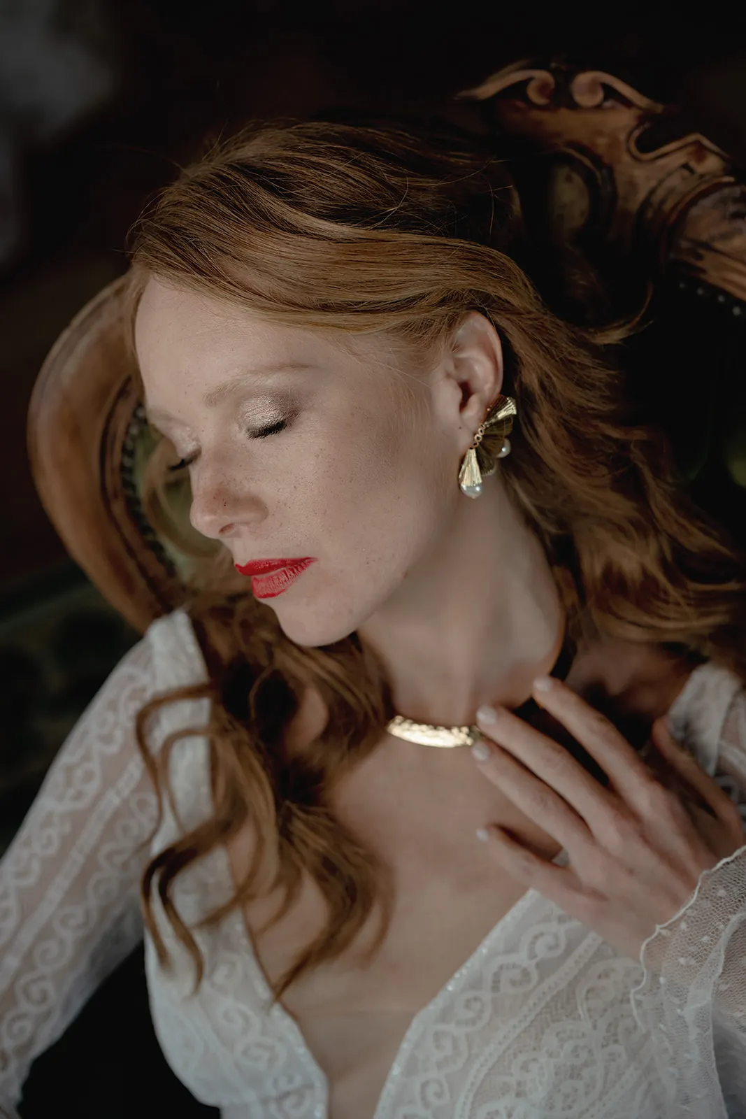 A woman with fair skin and red hair sits with her eyes closed, wearing bold red lipstick, gold earrings, and a gold necklace. Dressed in a white lace top, she gently touches her necklace—a serene portrait by a wedding photographer Tuscany. Tuscan Wedding Photographer