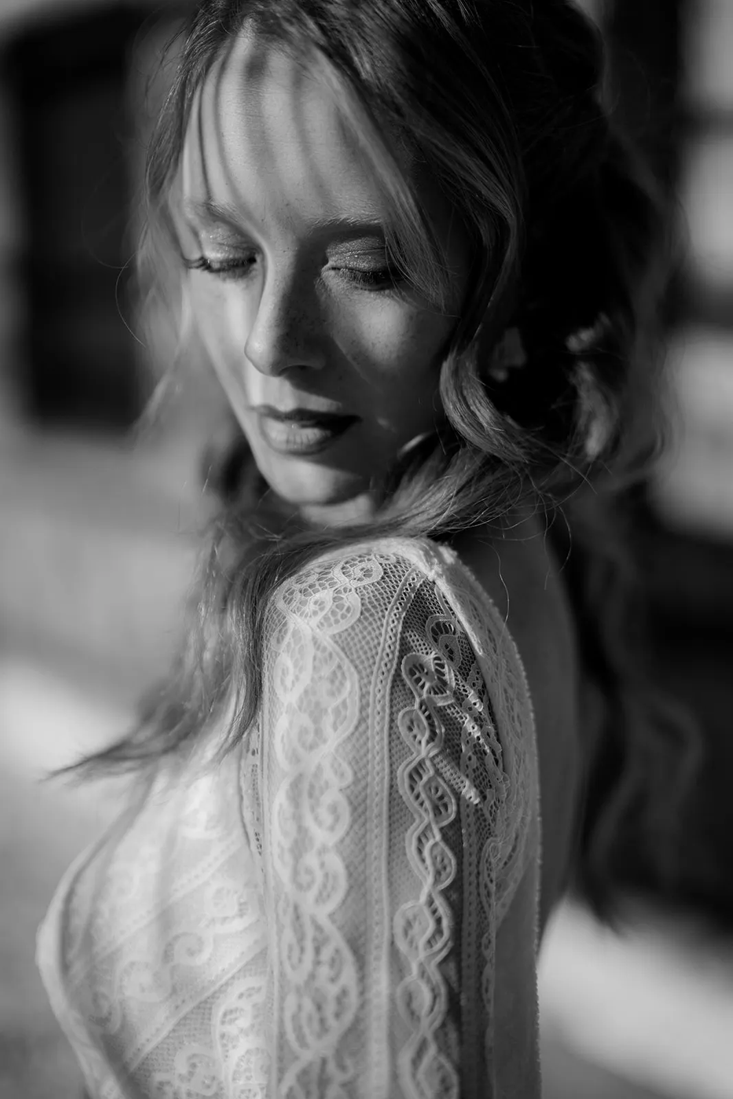 A woman in a lace dress stands outside, eyes closed, facing slightly down. Sunlight creates striped shadows across her face and shoulder—a timeless moment captured by a wedding photographer Tuscany style, with softly blurred black and white tones. Tuscan Wedding Photographer
