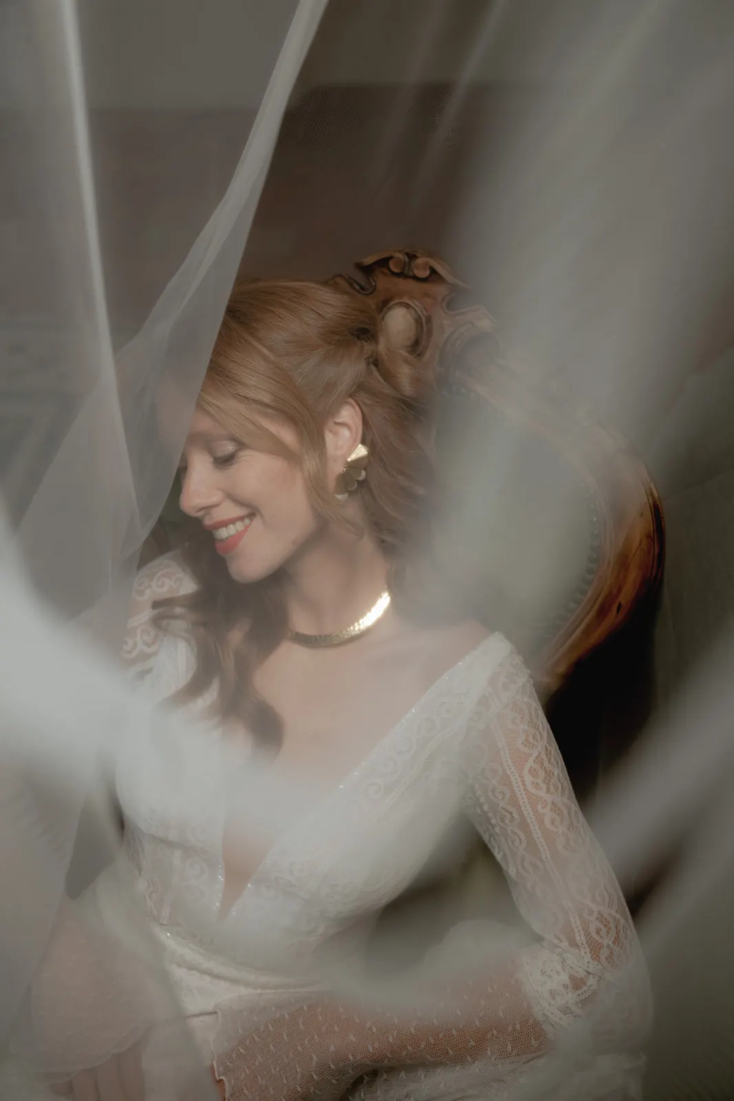 A woman with red hair in loose waves smiles with her eyes closed. Captured by a wedding photographer in Tuscany, she wears a white lace dress and gold earrings, sitting in a vintage chair, partially obscured by sheer white fabric for a dreamy, romantic effect. Tuscan Wedding Photographer