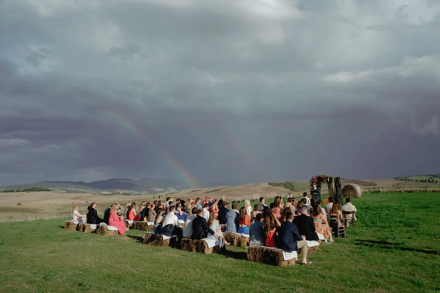 A group of people sits on hay bales at an outdoor wedding ceremony in Tuscany, facing a flower-adorned arch. The sky is cloudy with a faint double rainbow over rolling hills—perfect for a wedding photographer Tuscany dreams of. Tuscan Wedding Photographer