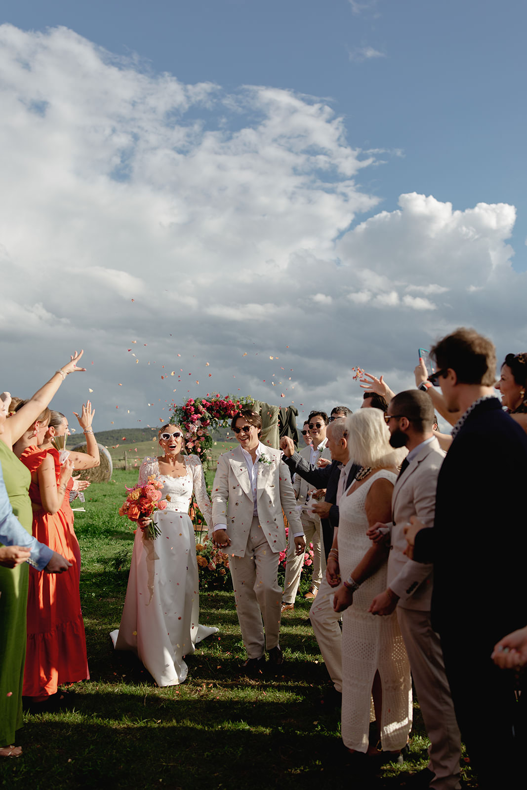 A bride and groom walk joyfully down an outdoor aisle in Tuscany, smiling as guests clap and toss flower petals. The bride holds a bouquet and wears sunglasses; a wedding photographer captures the festive moment beneath a colorful floral arch. Tuscan Wedding Photographer