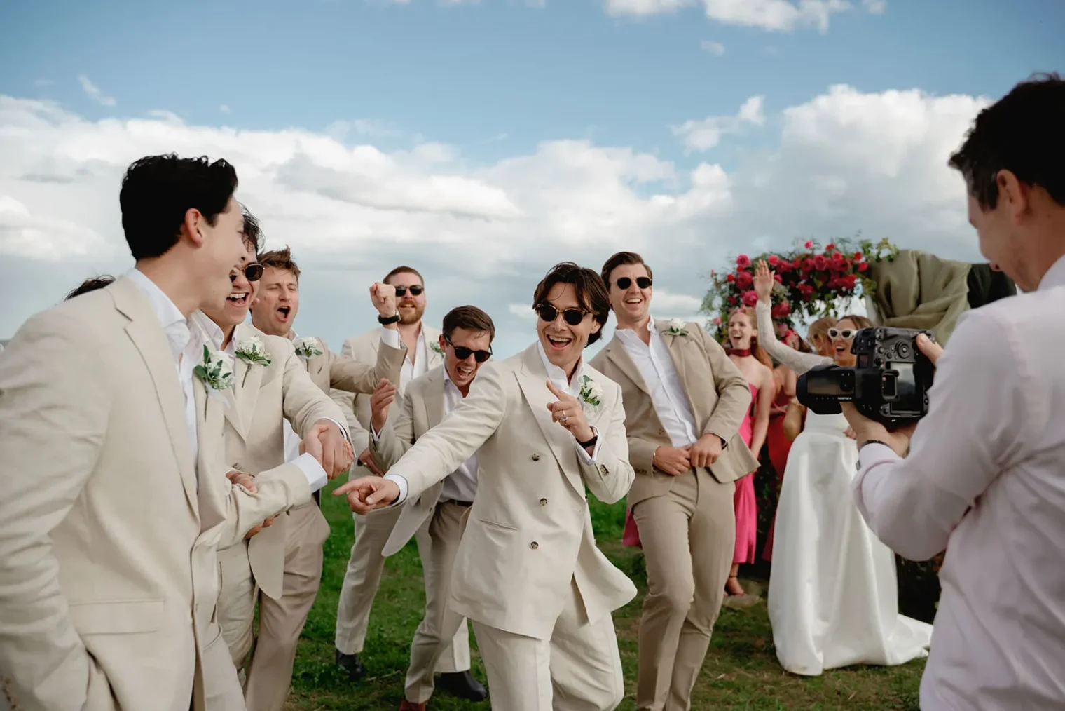 A joyful group of men in beige suits with sunglasses dance and laugh outdoors at a wedding in Tuscany. A wedding photographer captures the lively moment as women in pink dresses and a bride in white stand nearby under a floral arch beneath partly cloudy skies. Tuscan Wedding Photographer