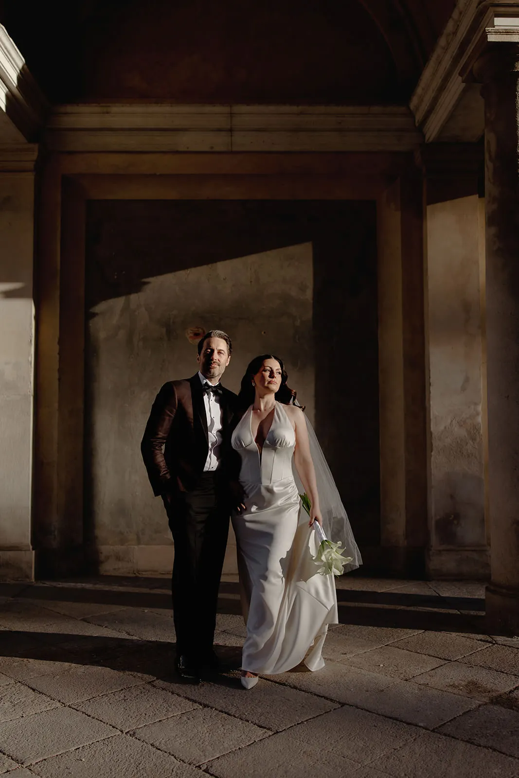 A bride in a white gown and veil stands arm-in-arm with a groom in a dark suit and bow tie, both smiling. Captured by a wedding photographer Tuscany, they stand under an archway with sunlight casting shadows on the stone floor behind them. Tuscan Wedding Photographer