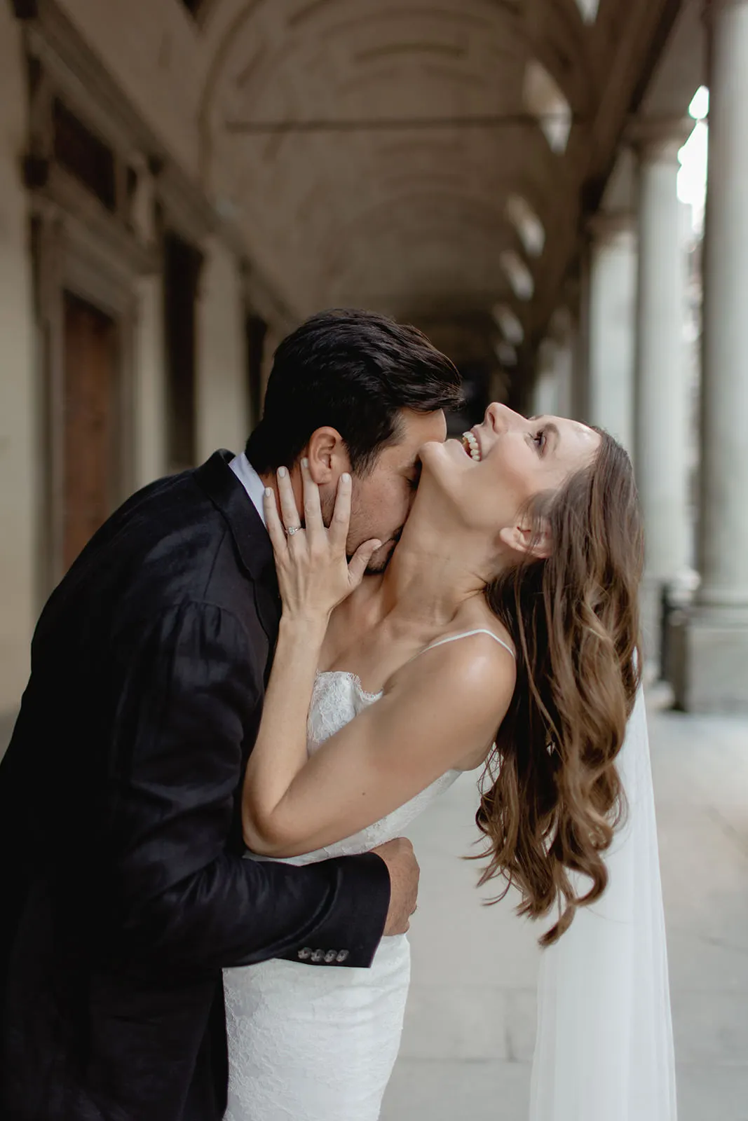 A groom in a black suit embraces and kisses a smiling bride in a white dress and veil under an elegant, arched corridor—captured perfectly by a wedding photographer Tuscany style as laughter and love fill the scene. Tuscan Wedding Photographer