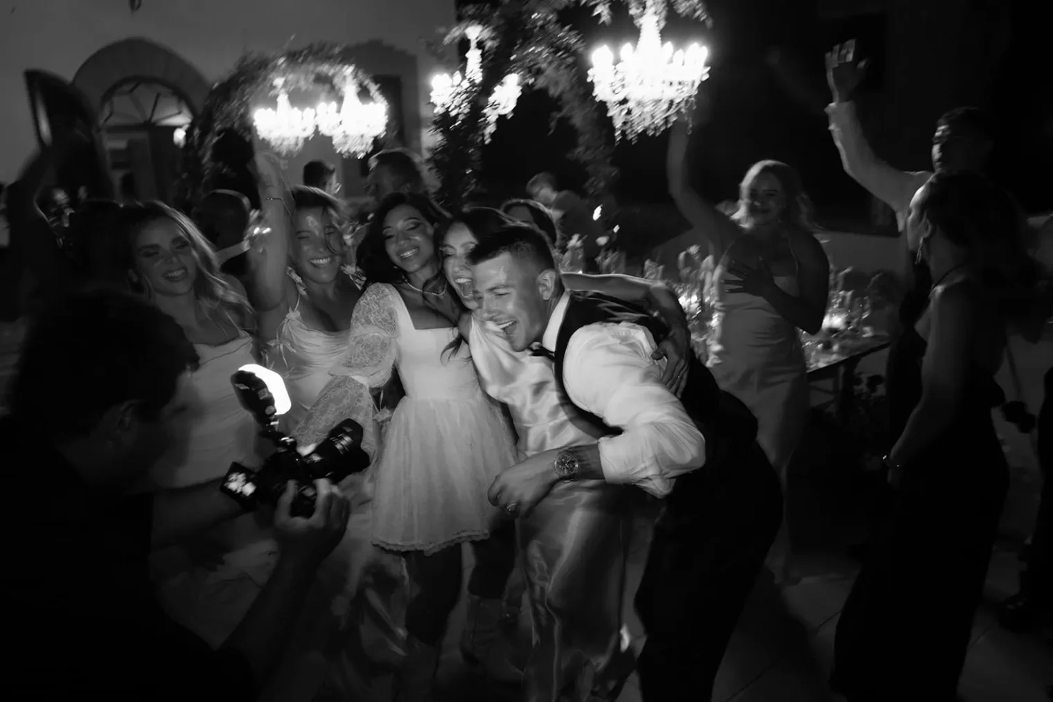 A joyful group dances and poses at a lively indoor party. In the foreground, a man and woman smile, embracing for a photo by a skilled wedding photographer Tuscany, as others cheer around them beneath elegant chandeliers and warm lights in the energetic atmosphere. Tuscan Wedding Photographer