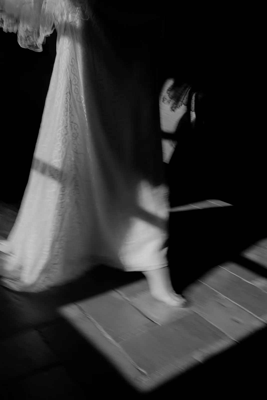 Black and white photo by a wedding photographer Tuscany: a person in a lace-trimmed dress walks barefoot on a wooden floor, sunlight casting geometric shadows. Only the lower half and hand are visible. Tuscan Wedding Photographer